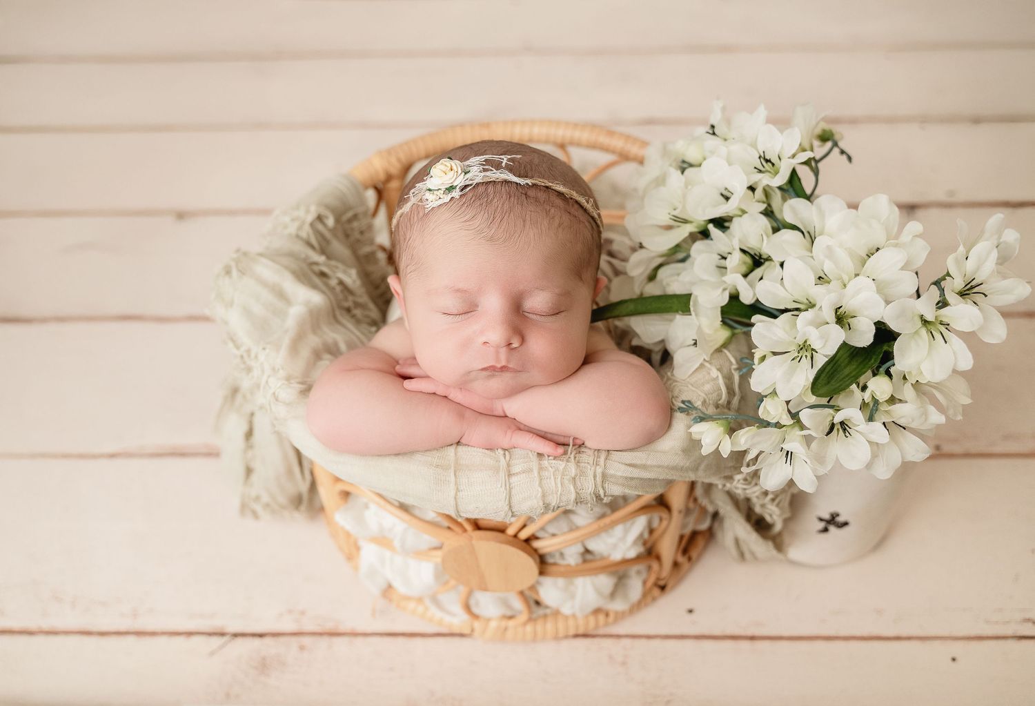 A sleeping newborn rests in a round basket with white lilies and vintage decor on wooden floorboards.