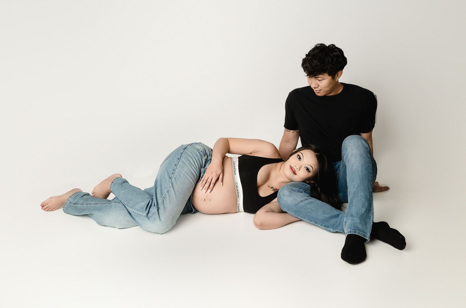 Minimalist maternity session on white background with casual denim and black attire.