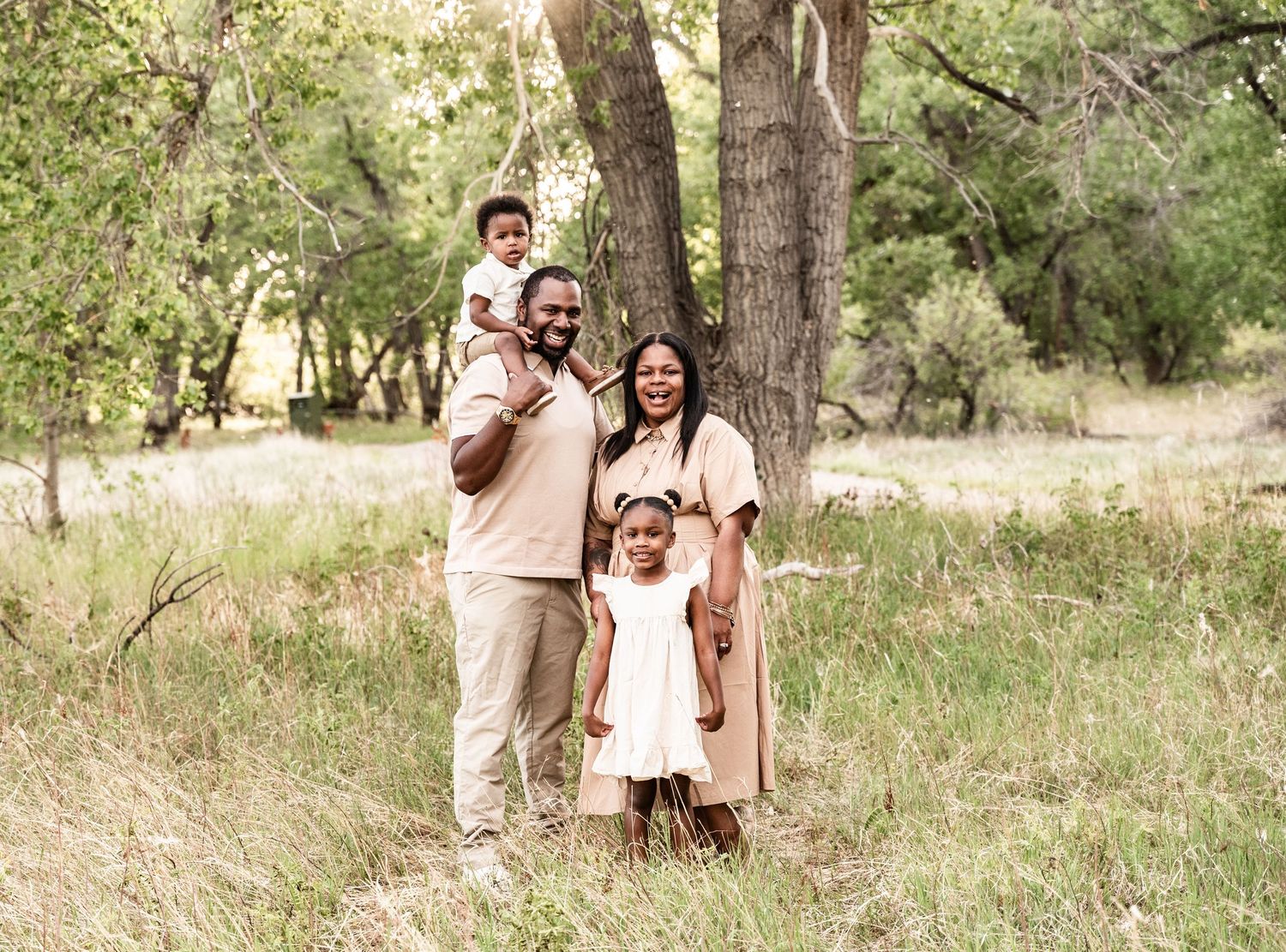 Happy Black family of four posing outdoors in a wooded field, wearing coordinated beige outfits for a portrait.