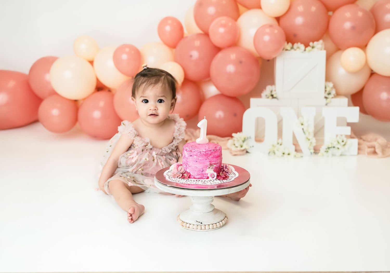 Baby's first birthday celebration with pink cake and coral balloon backdrop.