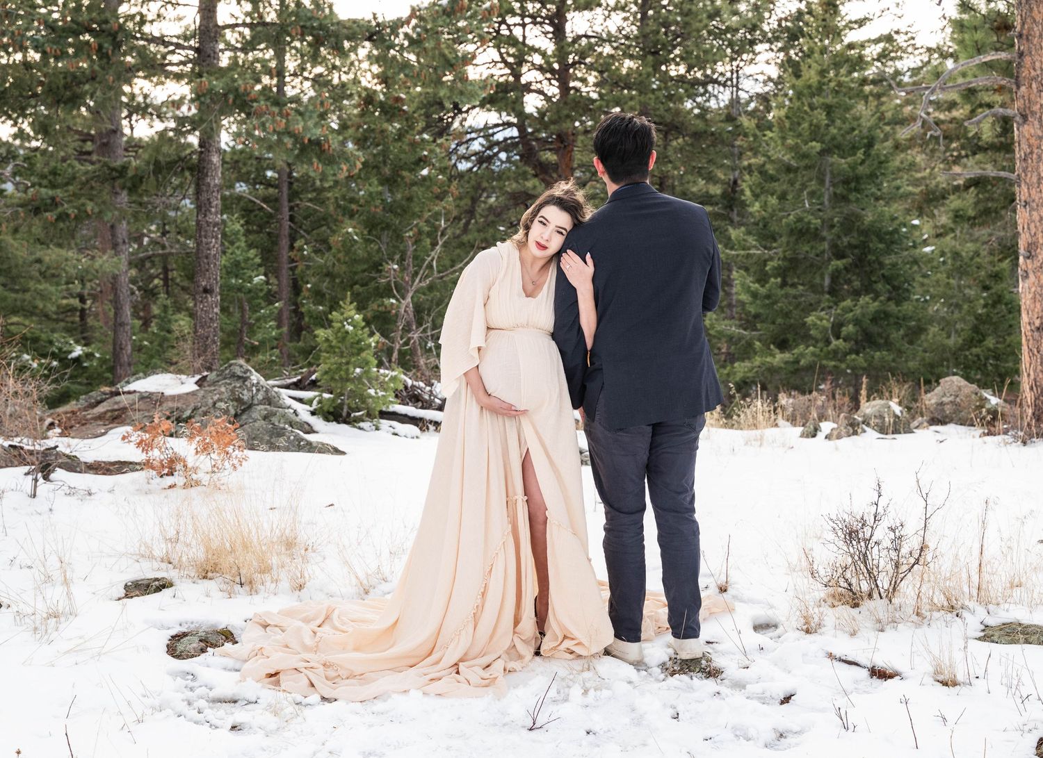 A romantic winter photoshoot in a snowy forest with evergreen trees and soft peach-colored dress details.