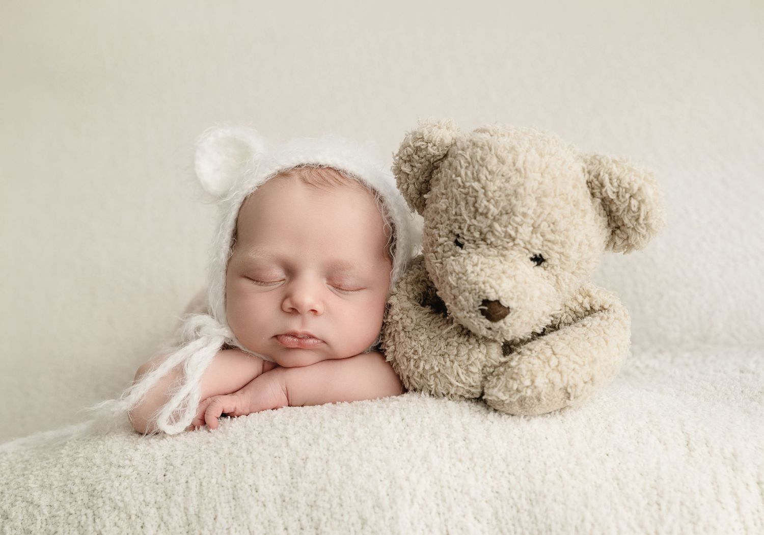 Newborn baby sleeping peacefully in white bear bonnet beside a soft teddy bear on a fluffy cream blanket.