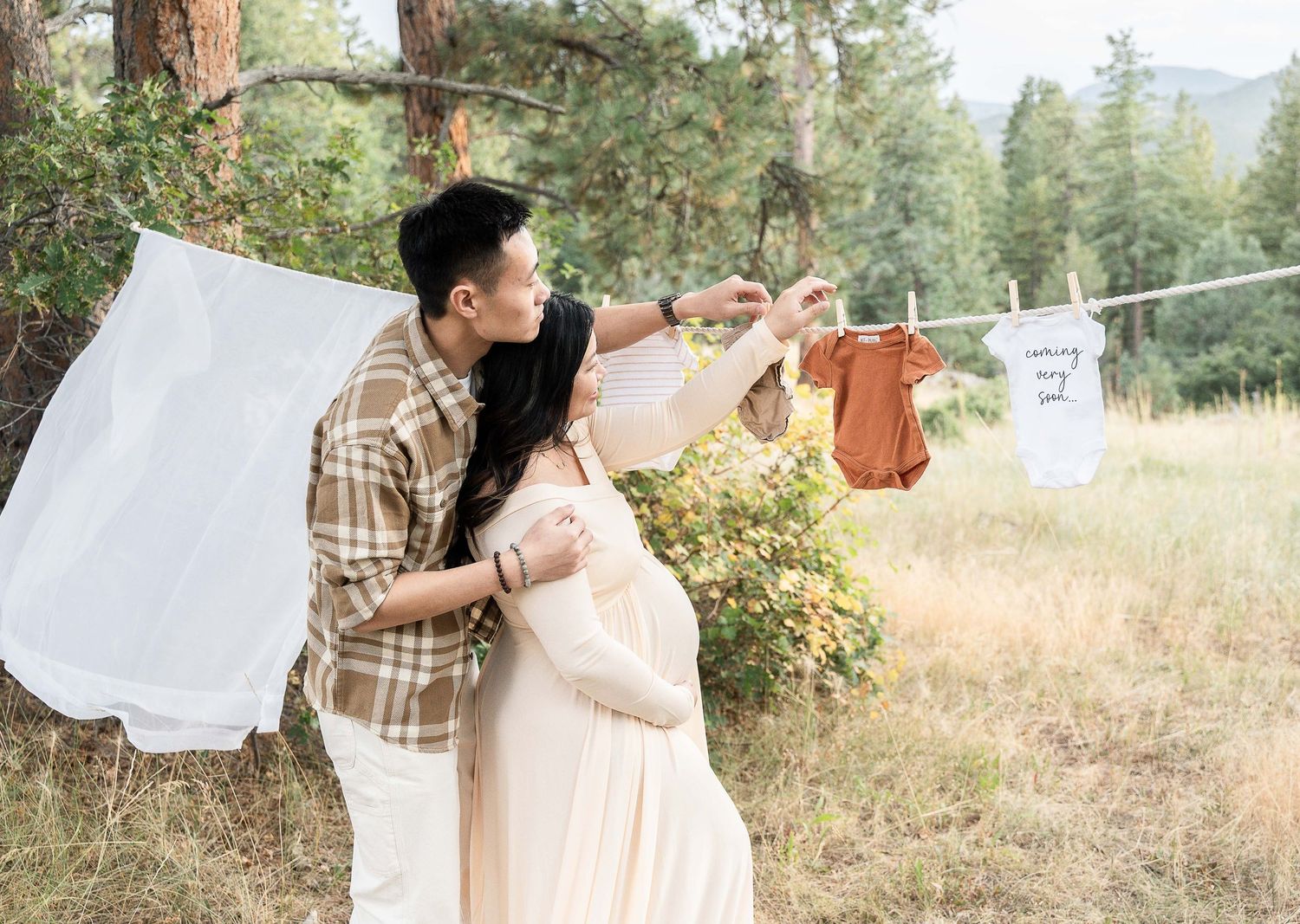 A couple in neutral tones embraces outdoors against a backdrop of tall pine trees and natural scenery.