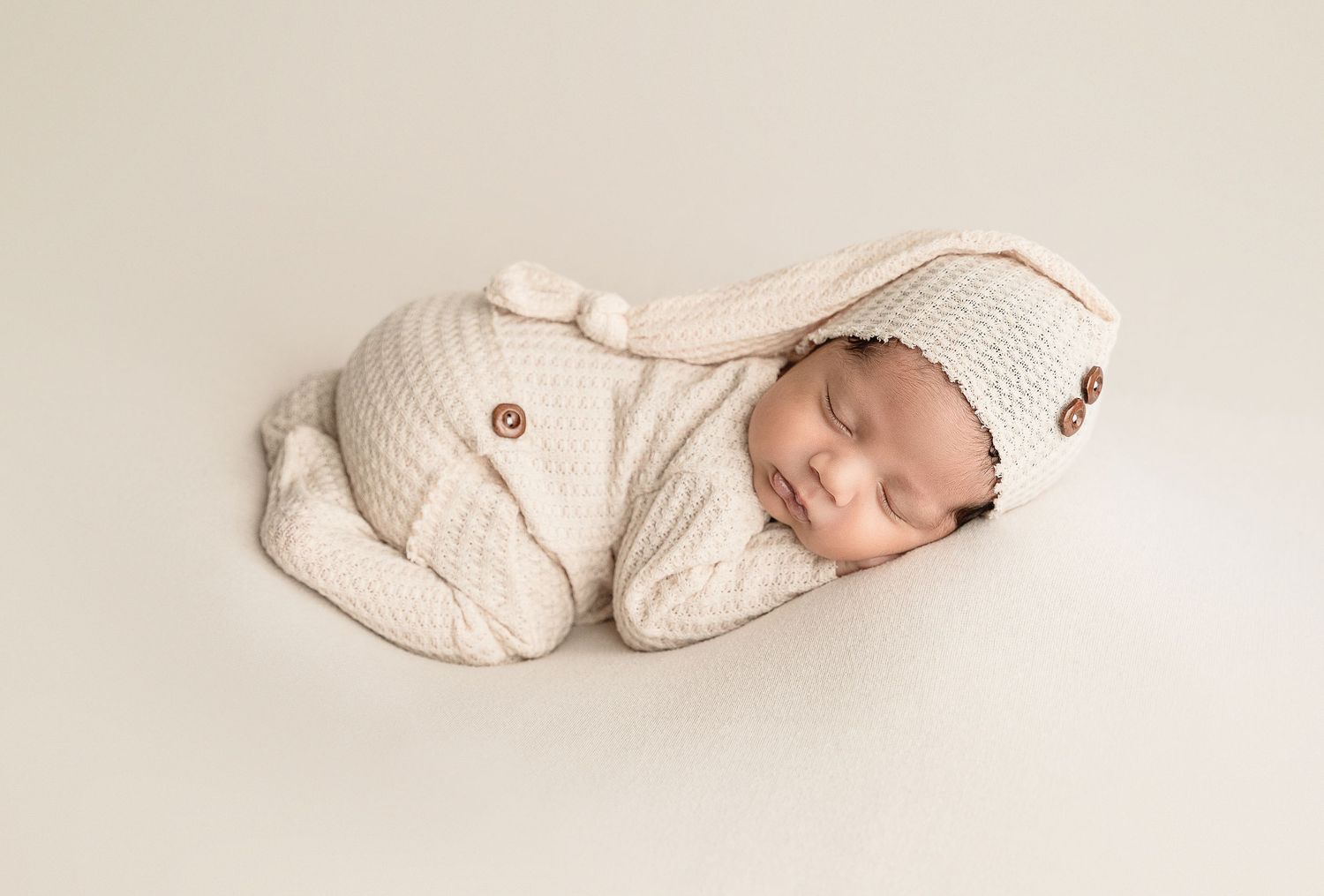 Sleeping newborn baby curled up in a cozy cream-colored knit sweater and hat on neutral backdrop.