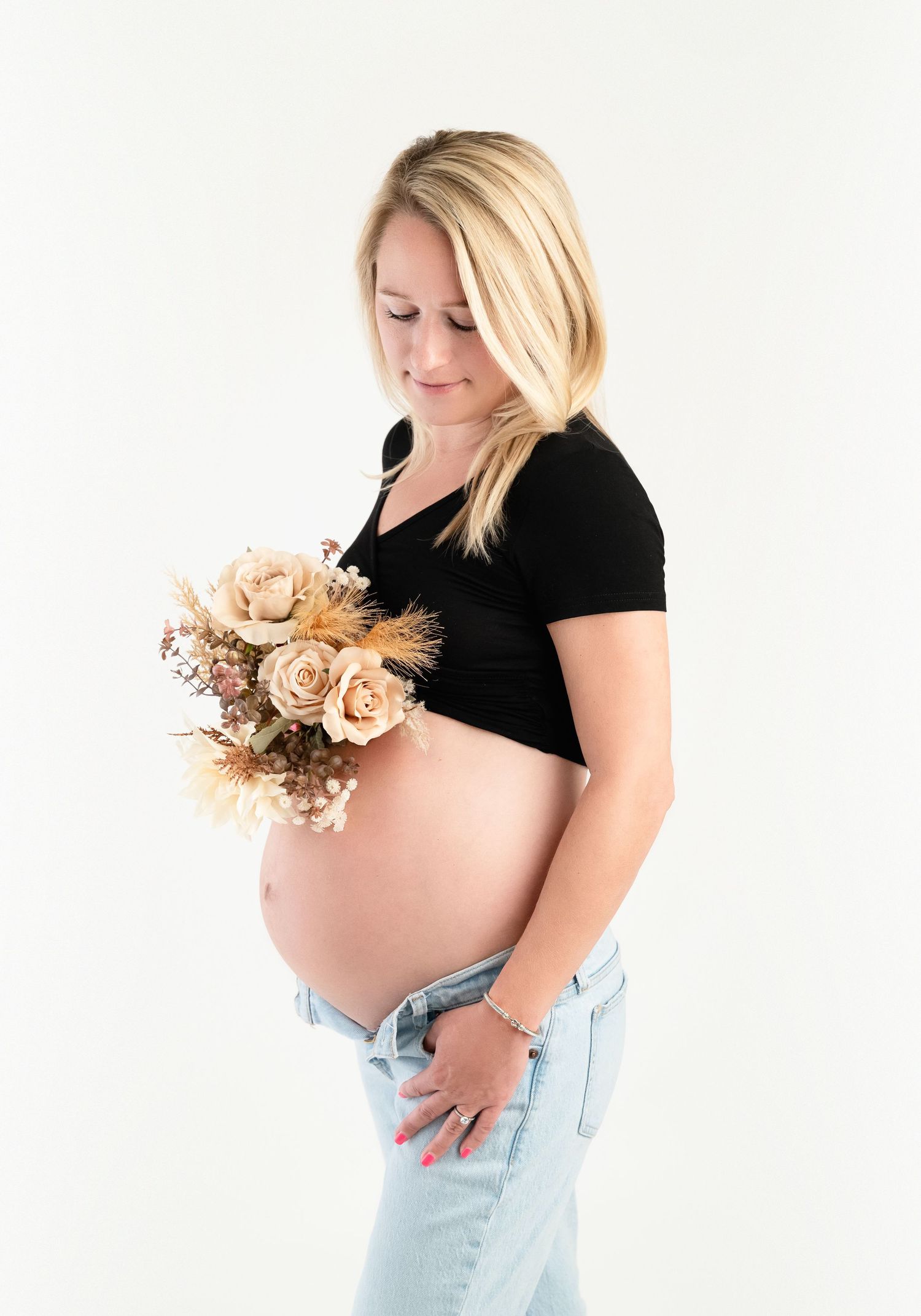 Pregnant woman in black crop top and jeans holding beige dried flower bouquet against white background.