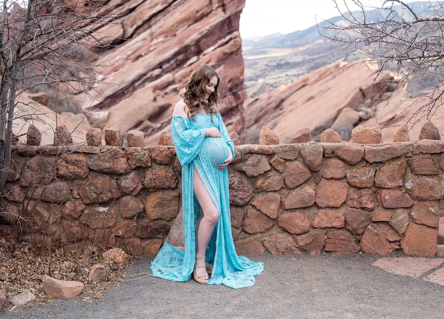 Someone in a flowing turquoise gown poses by a rustic stone wall with dramatic red rocks in the background at Red Rocks Park.