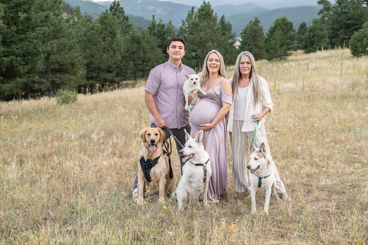Family group portrait with three dogs standing together in a mountain meadow with evergreen trees in the background.