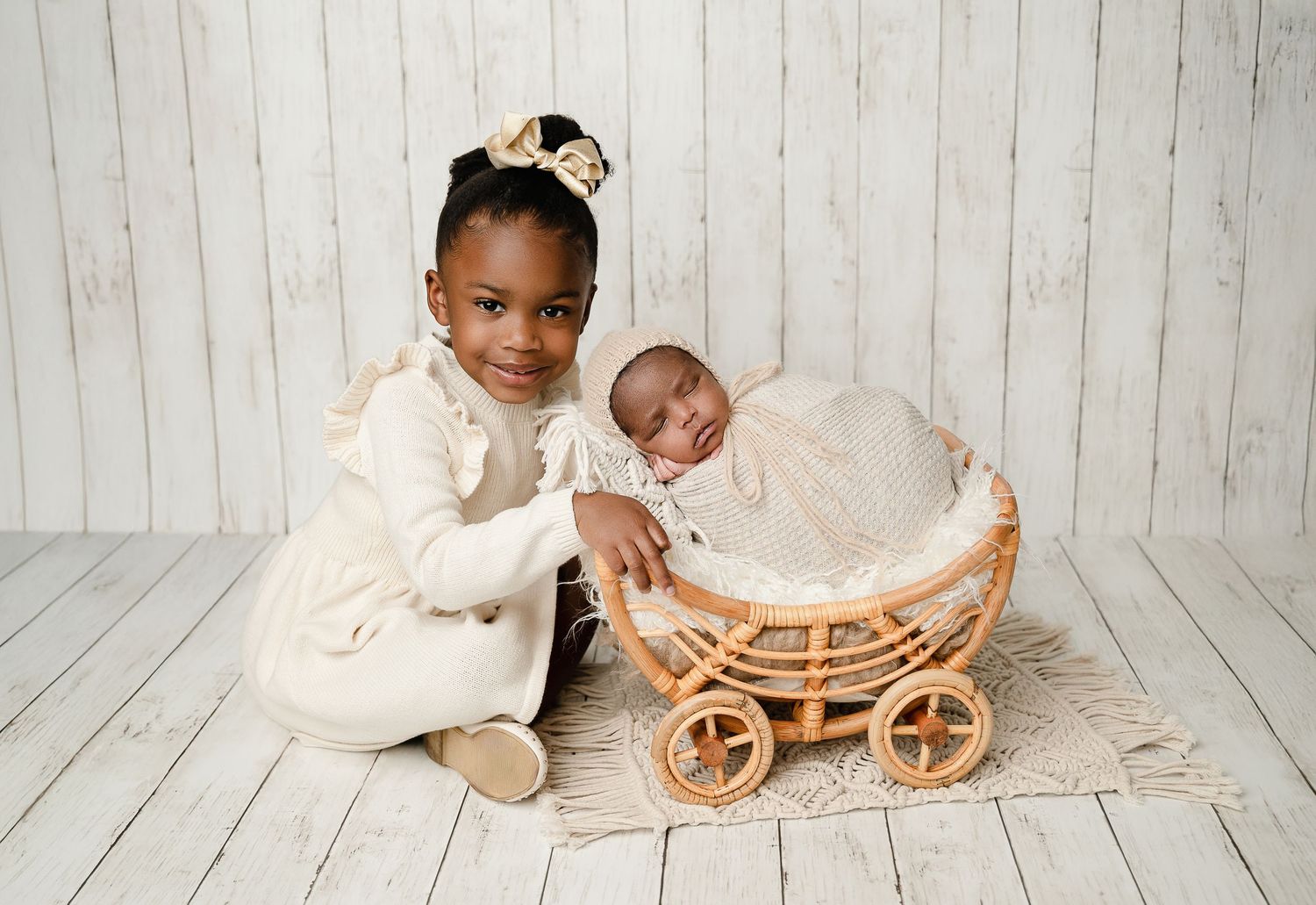 Two young children posing with decorative wicker carriage against white wooden backdrop.