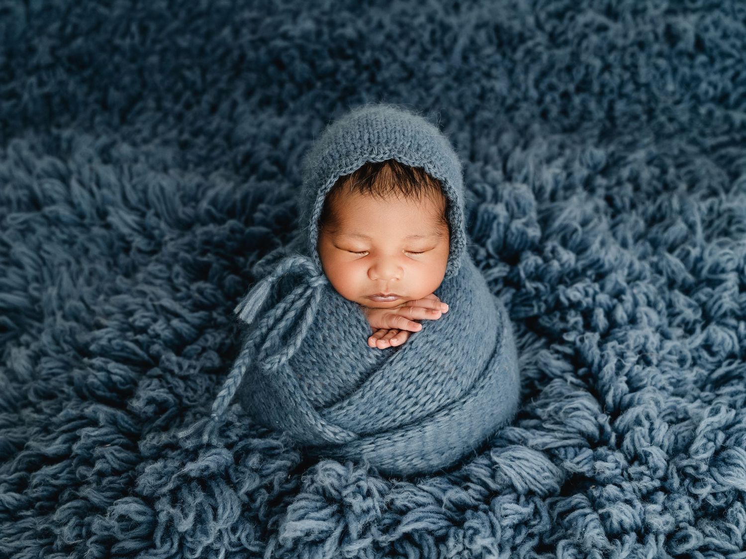 Newborn baby swaddled in blue knit wrap and bonnet, sleeping peacefully on a soft blue fluffy rug.