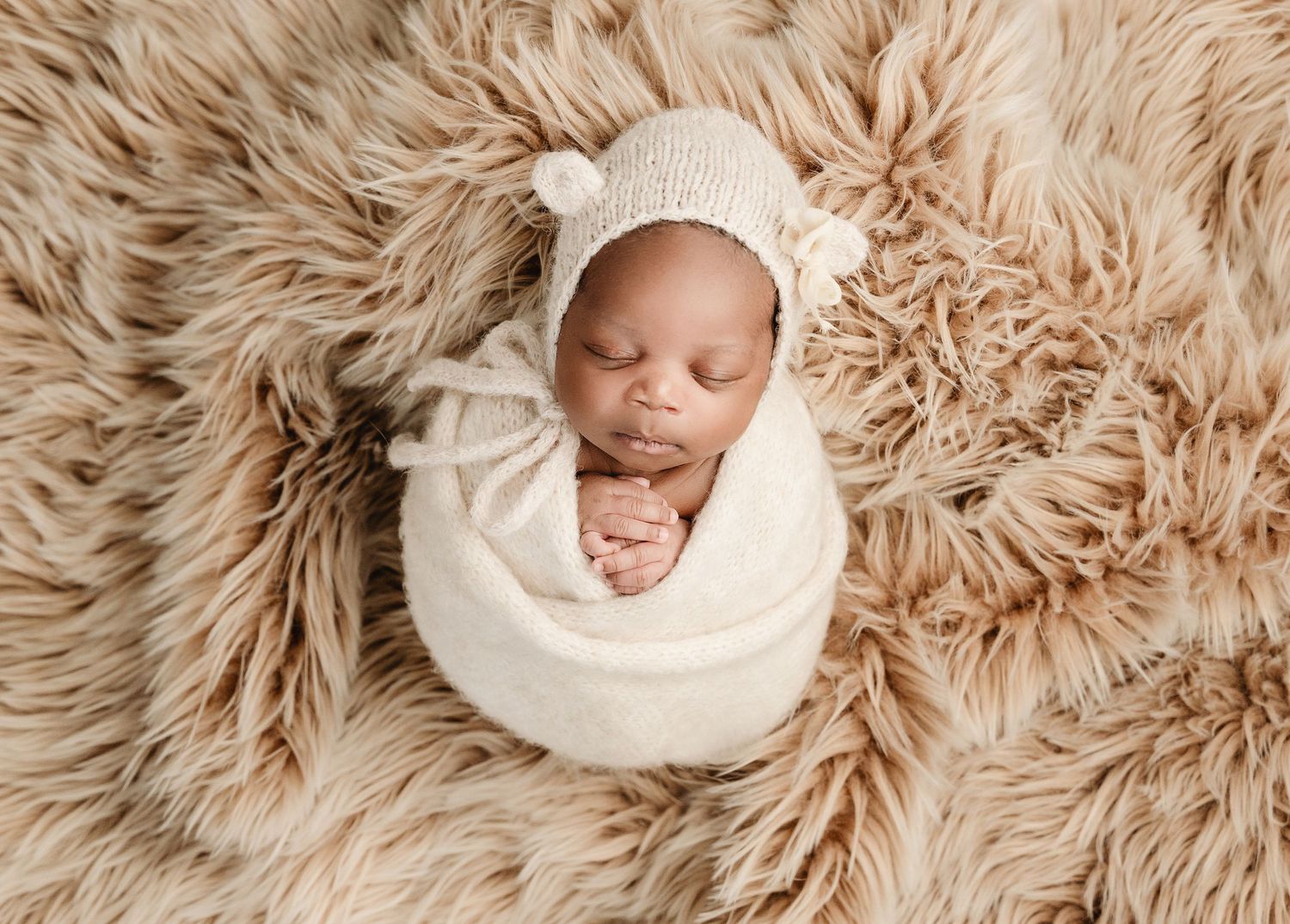 Newborn baby wrapped in cream swaddle wearing bear bonnet hat, sleeping peacefully on fluffy beige fur backdrop.