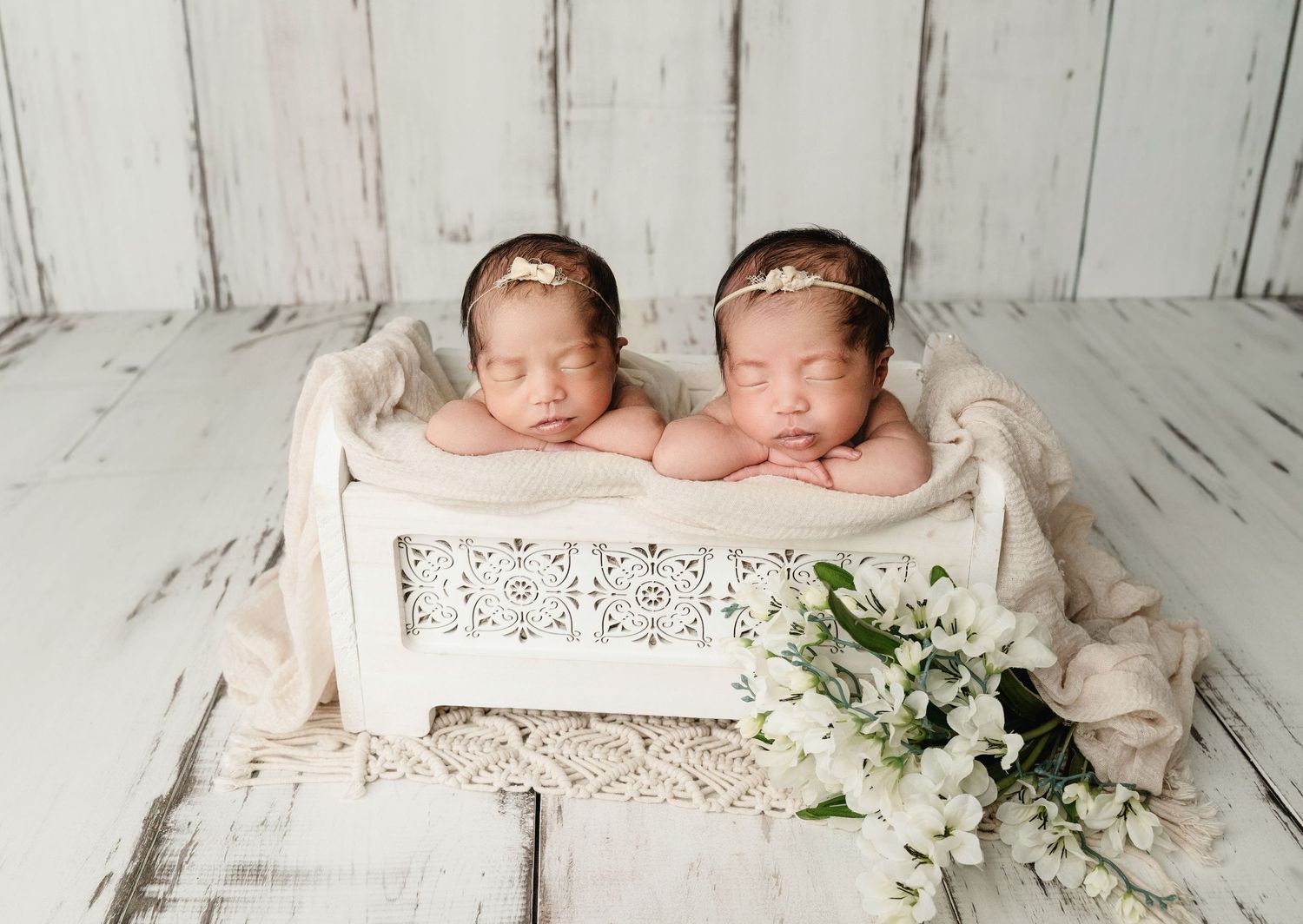 Two sleeping newborn twin babies in a white wooden bed with lace detail, adorned with gold headbands and white flowers.
