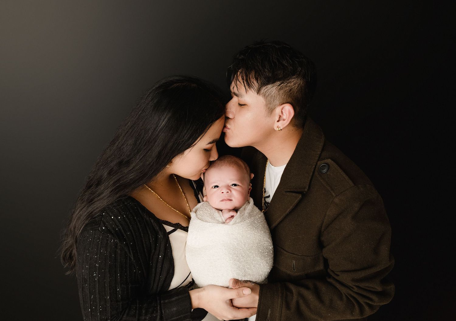 A loving couple embraces in a dimly lit setting while holding their baby in a tender family portrait.