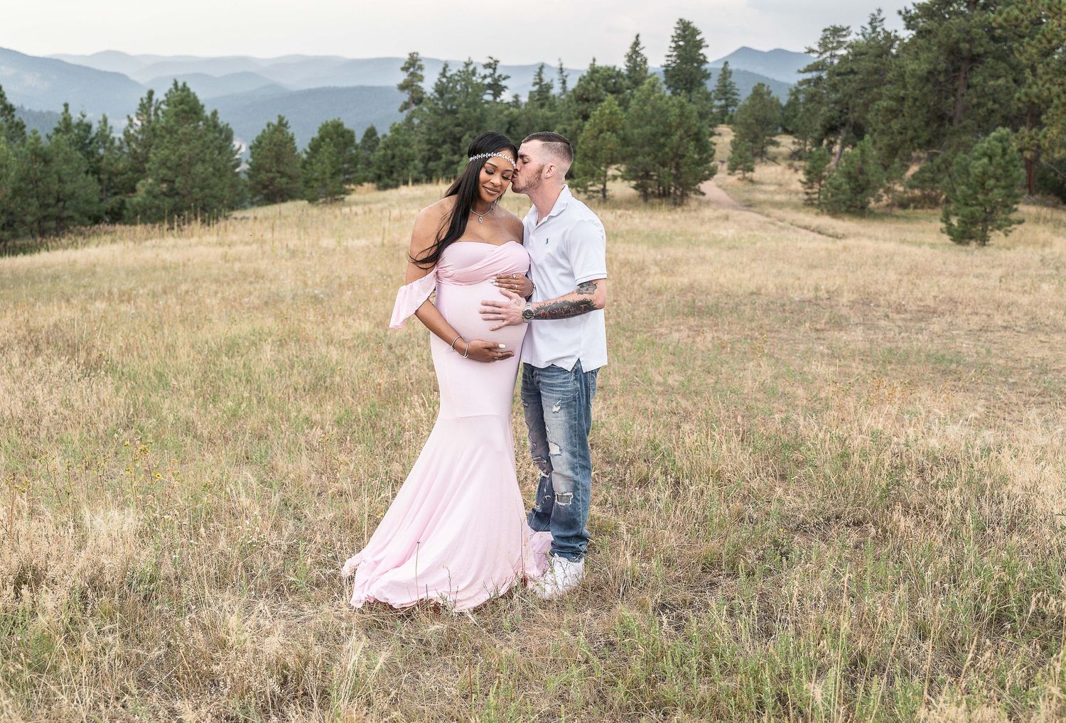 A couple shares romantic moments in a scenic mountain meadow wearing pink and white outfits during an engagement photoshoot.