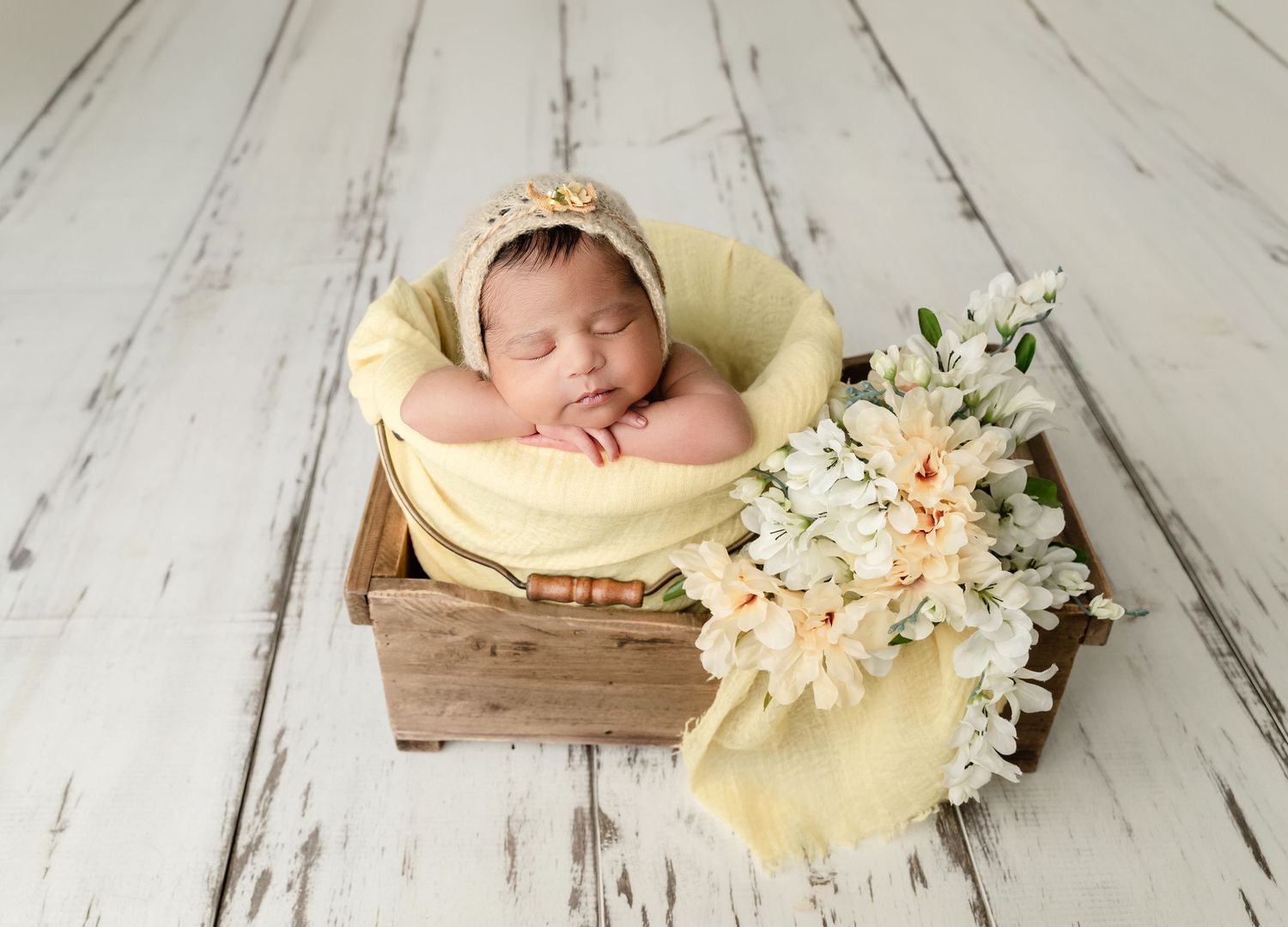 Sleeping newborn baby in yellow wrap and bonnet resting in wooden box with white flowers on rustic floor.