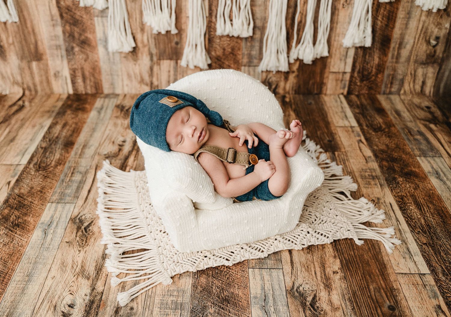 Sleeping infant curled up in blue knit outfit on white lace blanket against rustic wooden floor background.