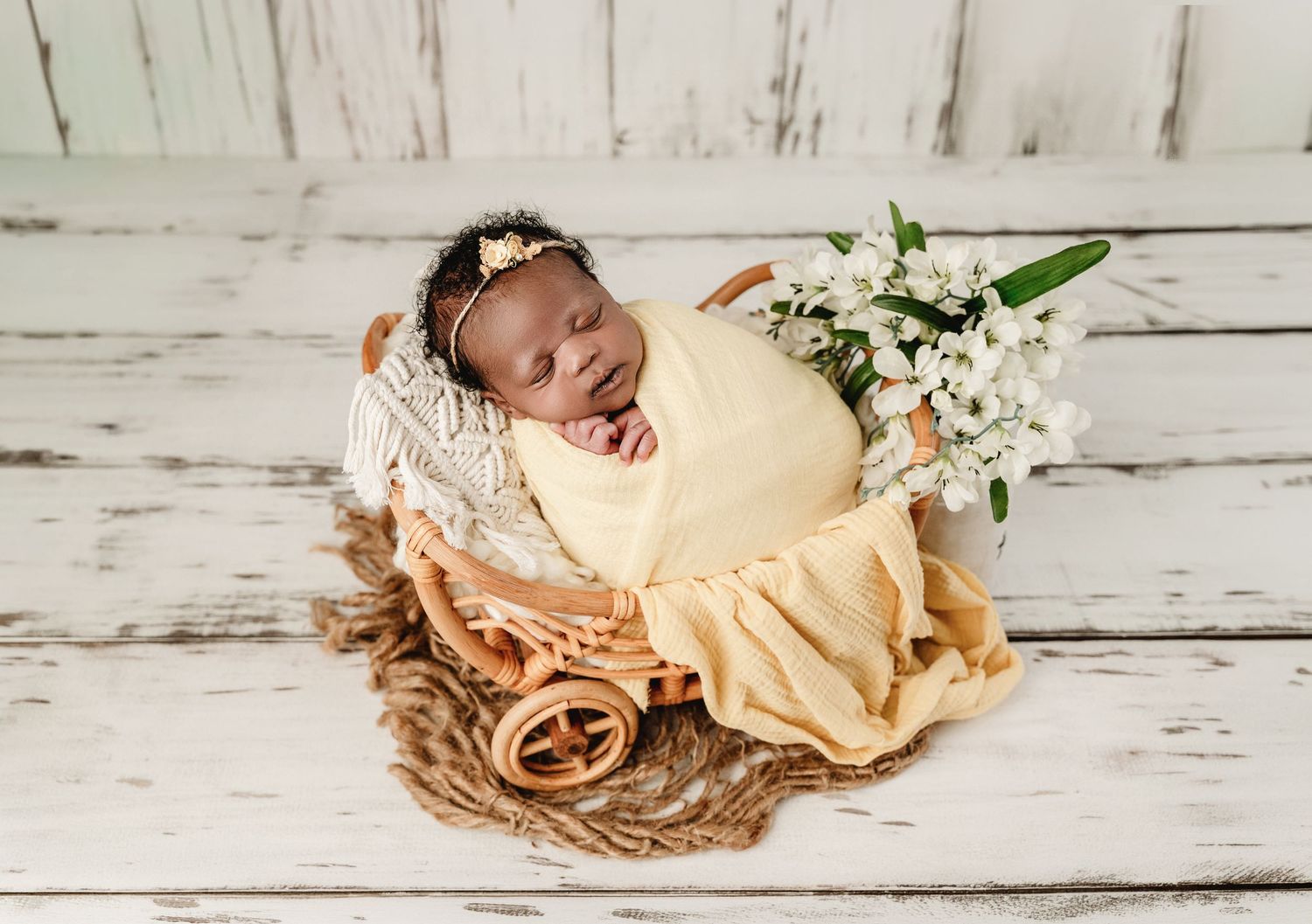 Newborn baby swaddled in yellow wrap sleeping in a wicker basket with white flowers on a rustic white wood background.