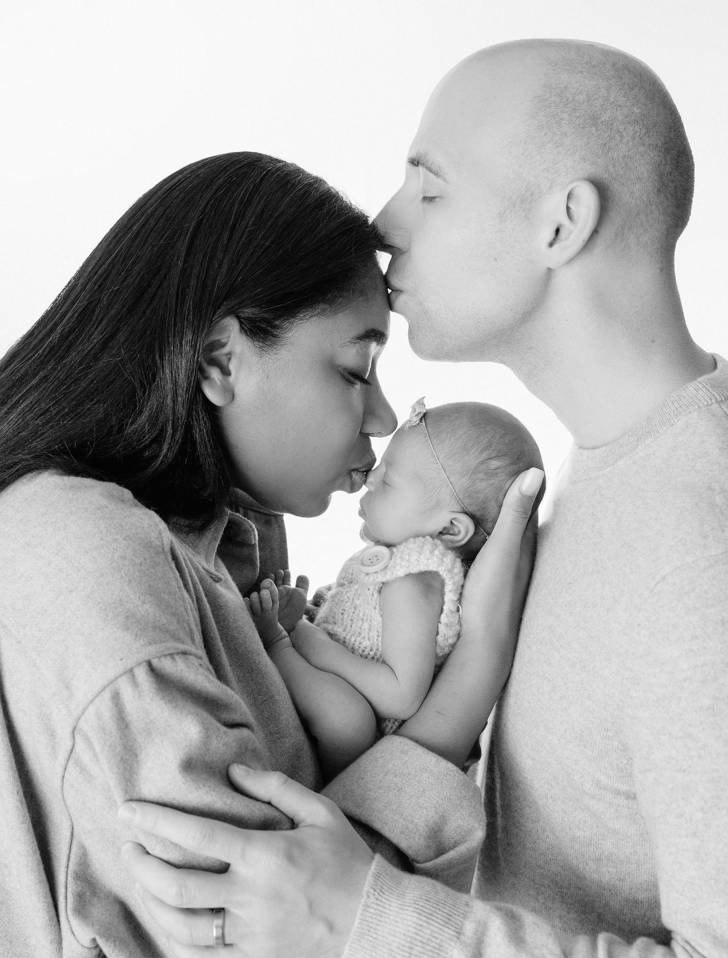 Black and white photo of parents kissing over their newborn baby, a tender family portrait moment.