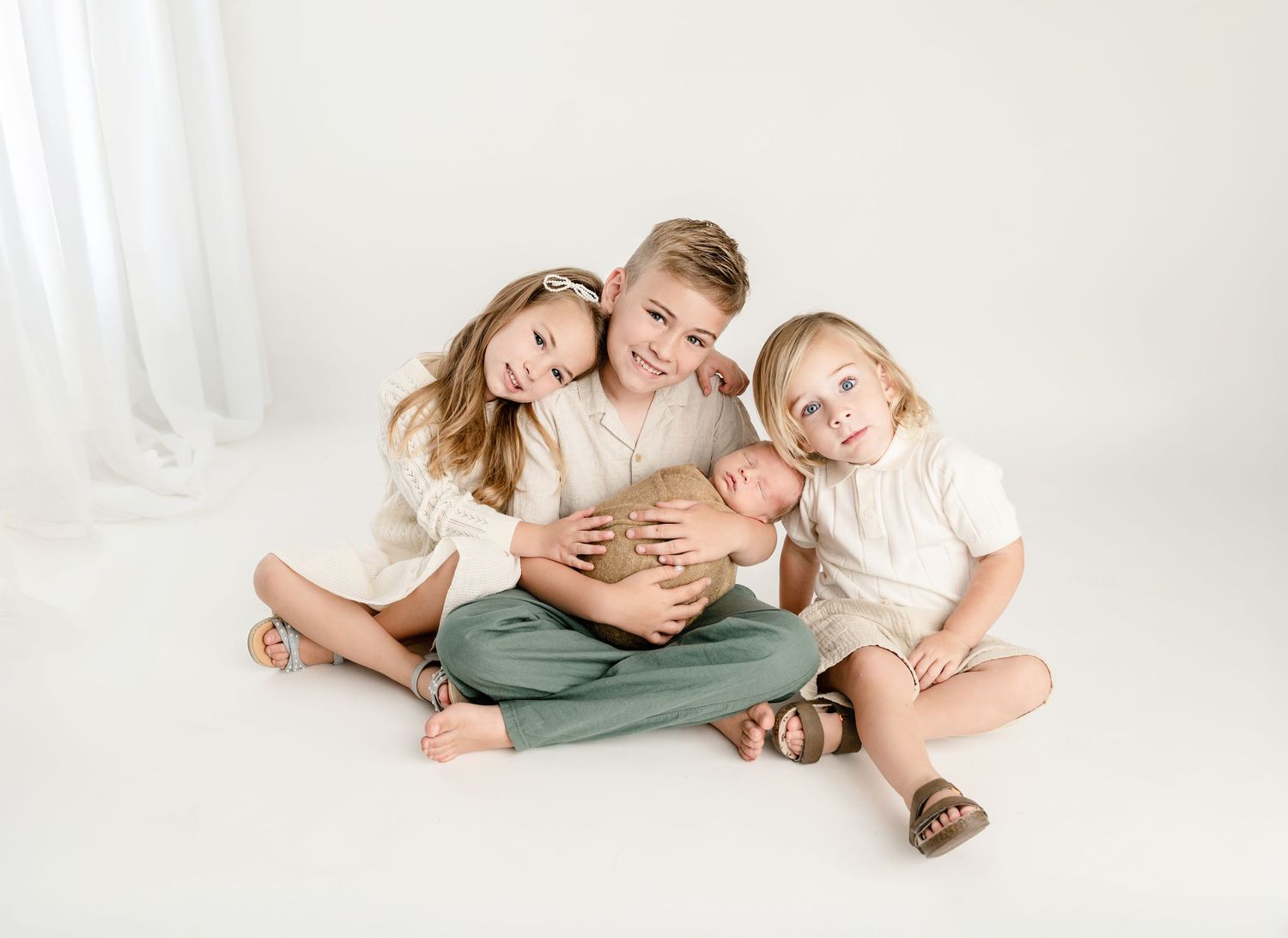Three siblings in cream outfits sit together on white backdrop, older kids hugging while holding a sleeping newborn baby.