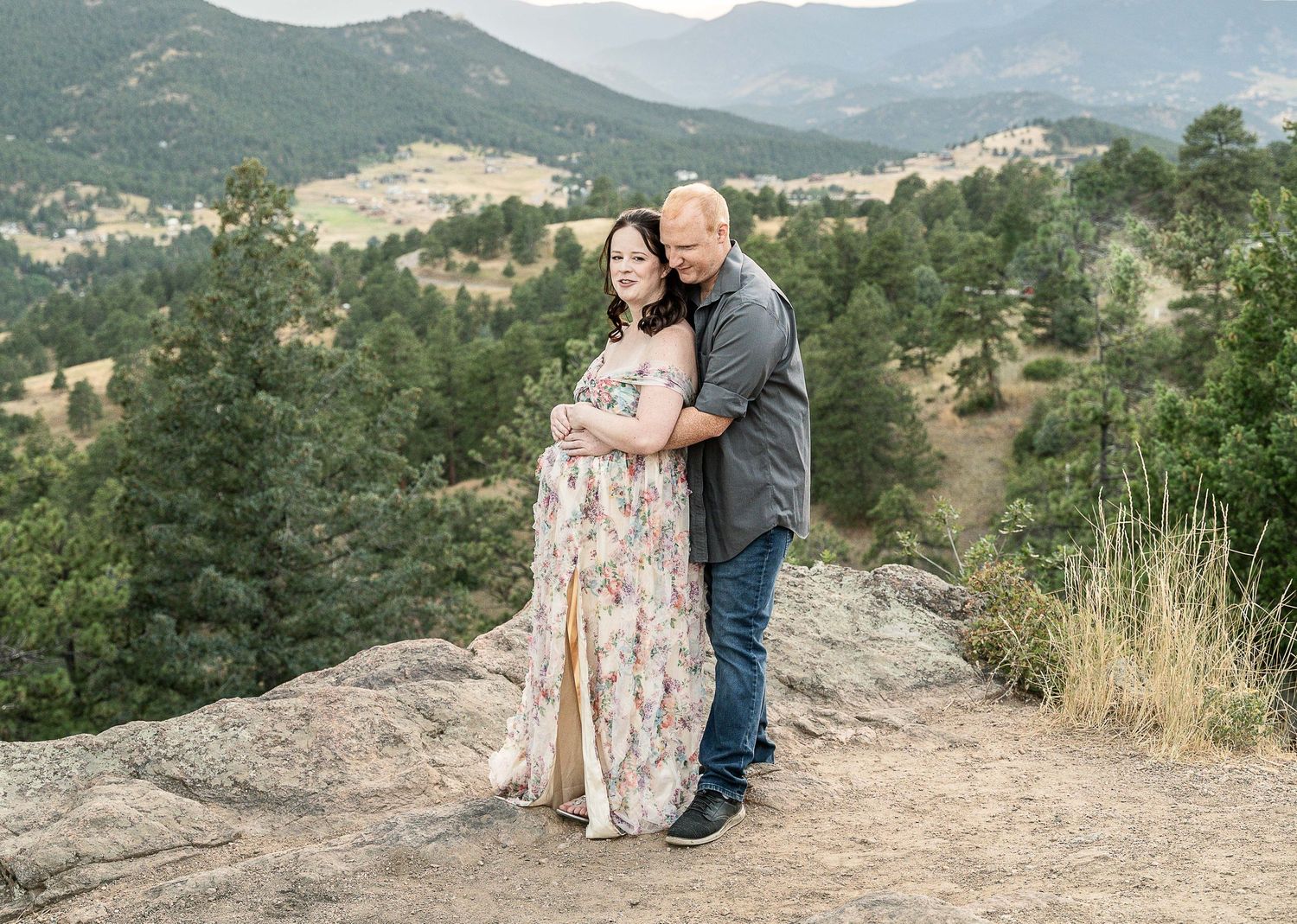 A couple embraces on a rocky overlook with scenic mountain views and evergreen forests in the background. Maternity session Denver, Colorado
