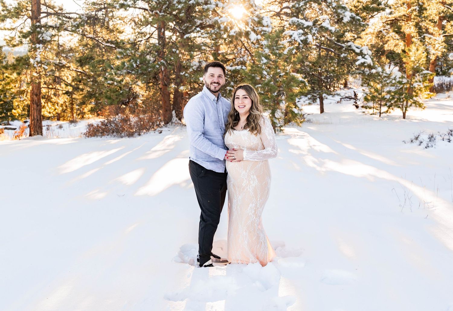 A couple embraces in a snowy winter landscape with golden sunlight filtering through the trees in the background.