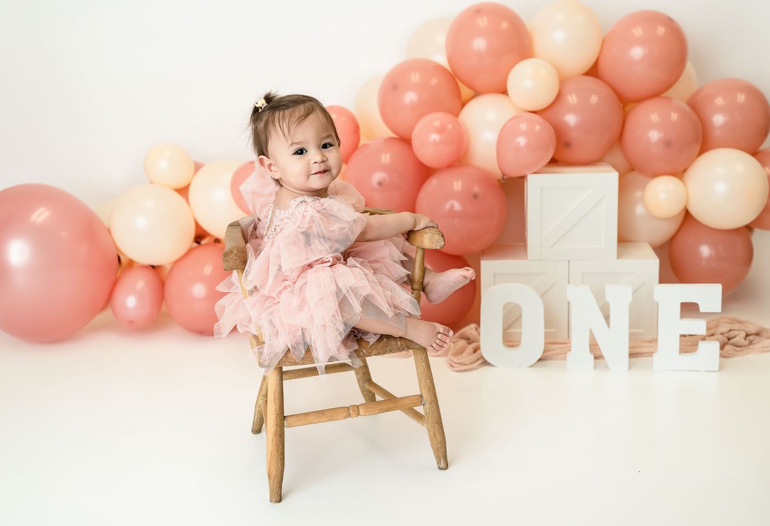 Baby's first birthday setup with pink balloons and rustic wooden chair against white backdrop.