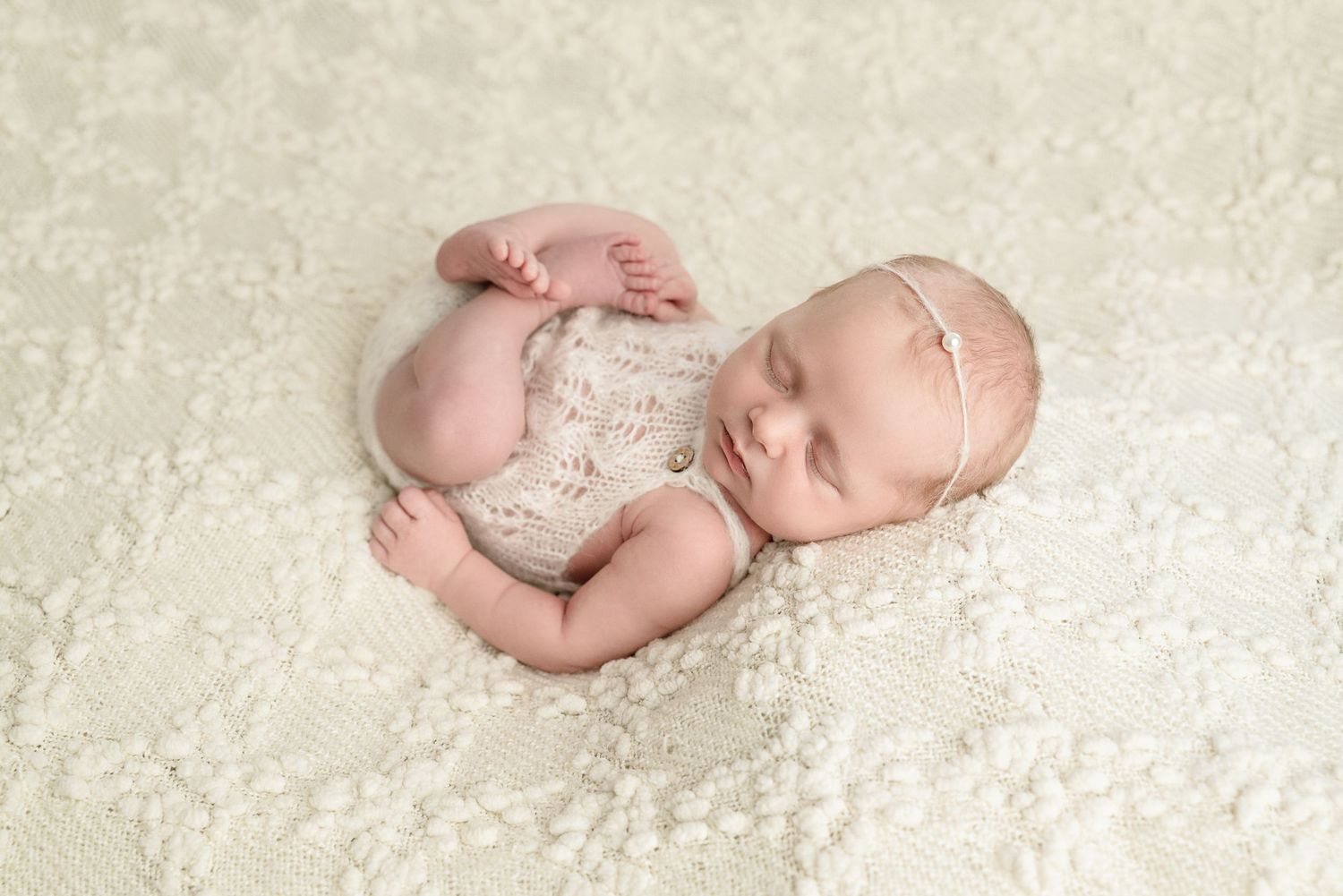 Sleeping newborn baby in cream lace romper and pink headband on white textured blanket.