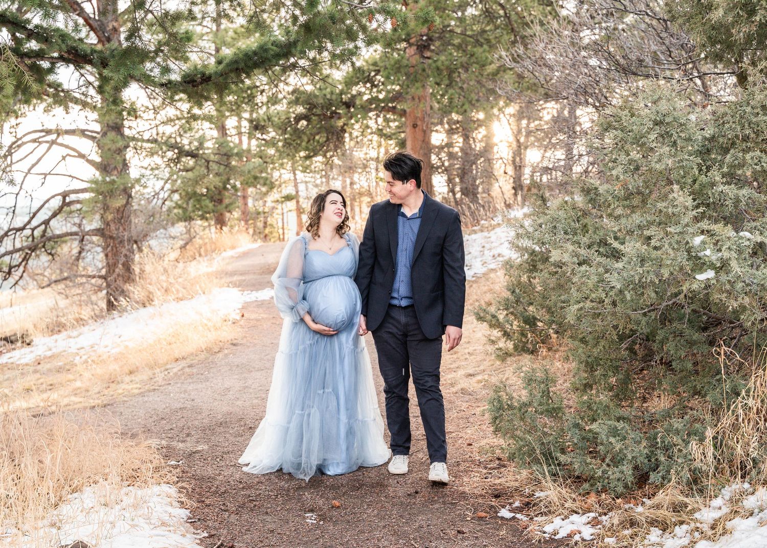 A couple walks hand in hand through a snowy winter forest path during golden hour.