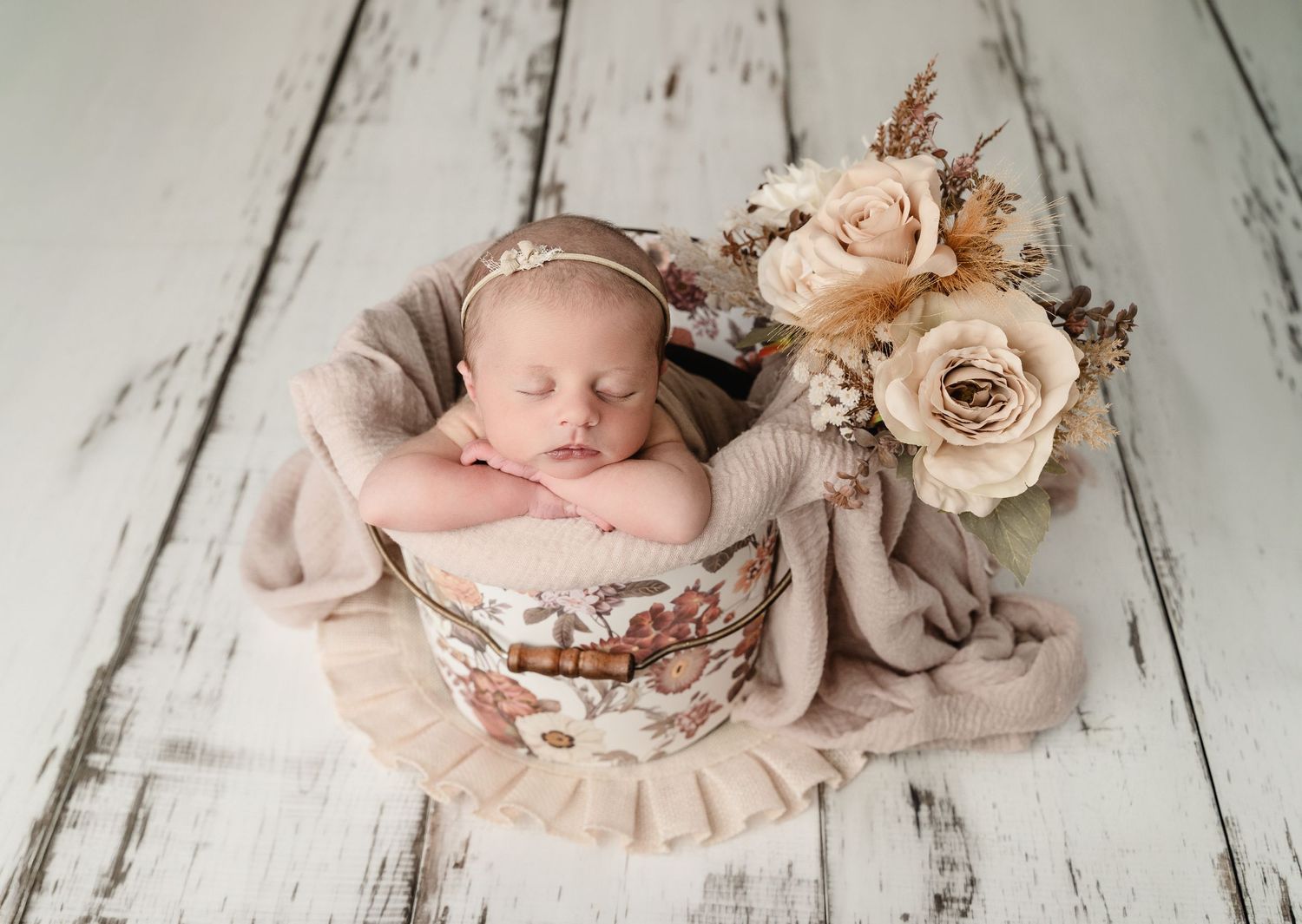 Newborn baby sleeping peacefully in a floral bucket with cream roses on a rustic white wood background.