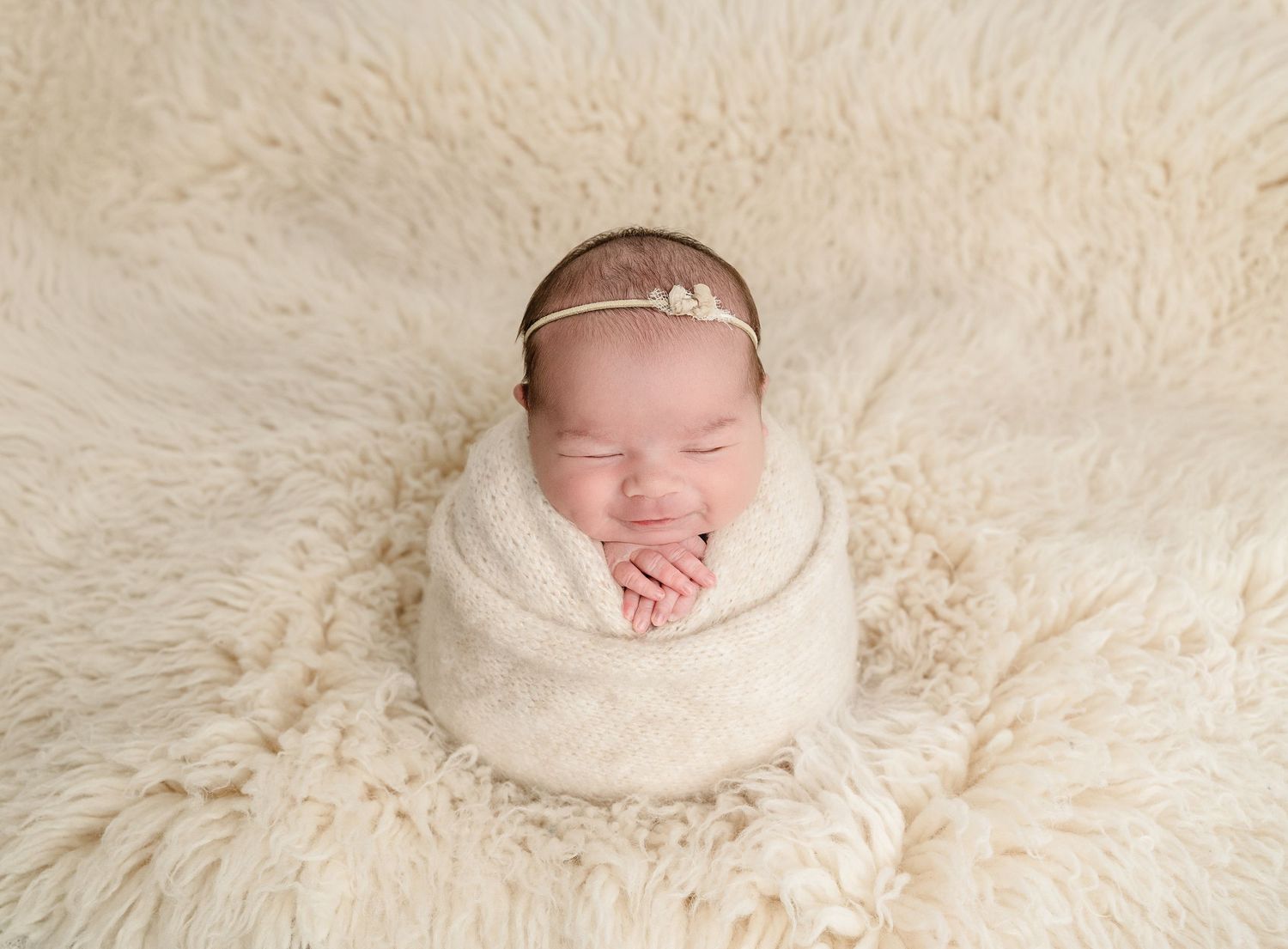 Sleeping newborn baby girl wrapped in cream knit swaddle with floral headband on fluffy white fur rug.