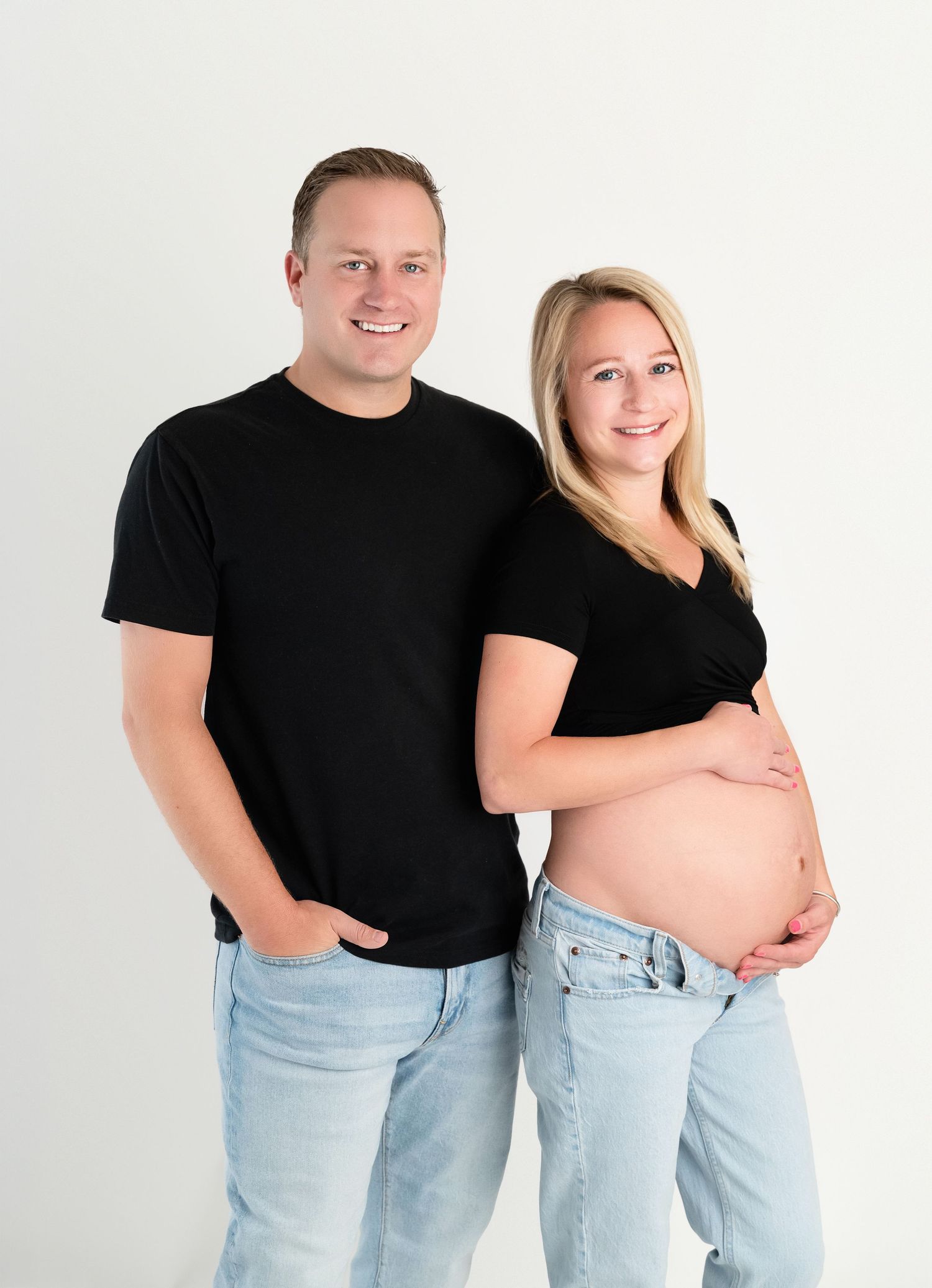 Smiling couple in matching black tops and jeans pose together, woman cradling pregnant belly, against white background.