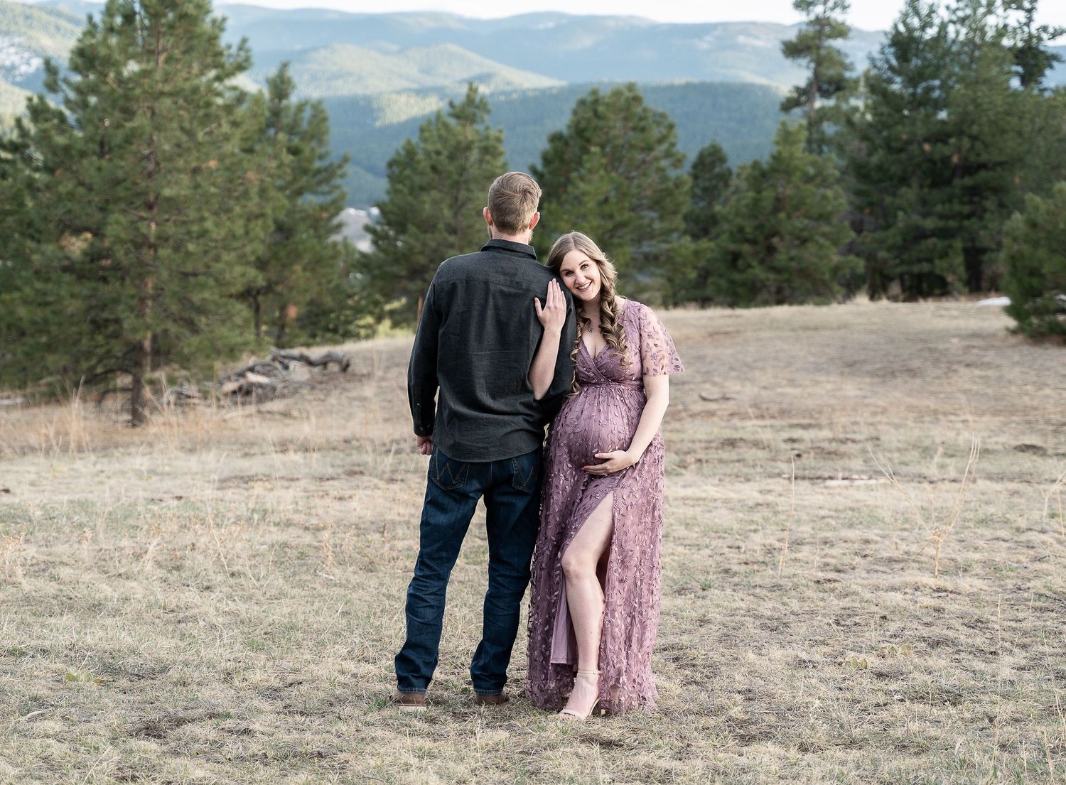 Couple walks together on grassy hillside with mountains and pine trees in the distance.