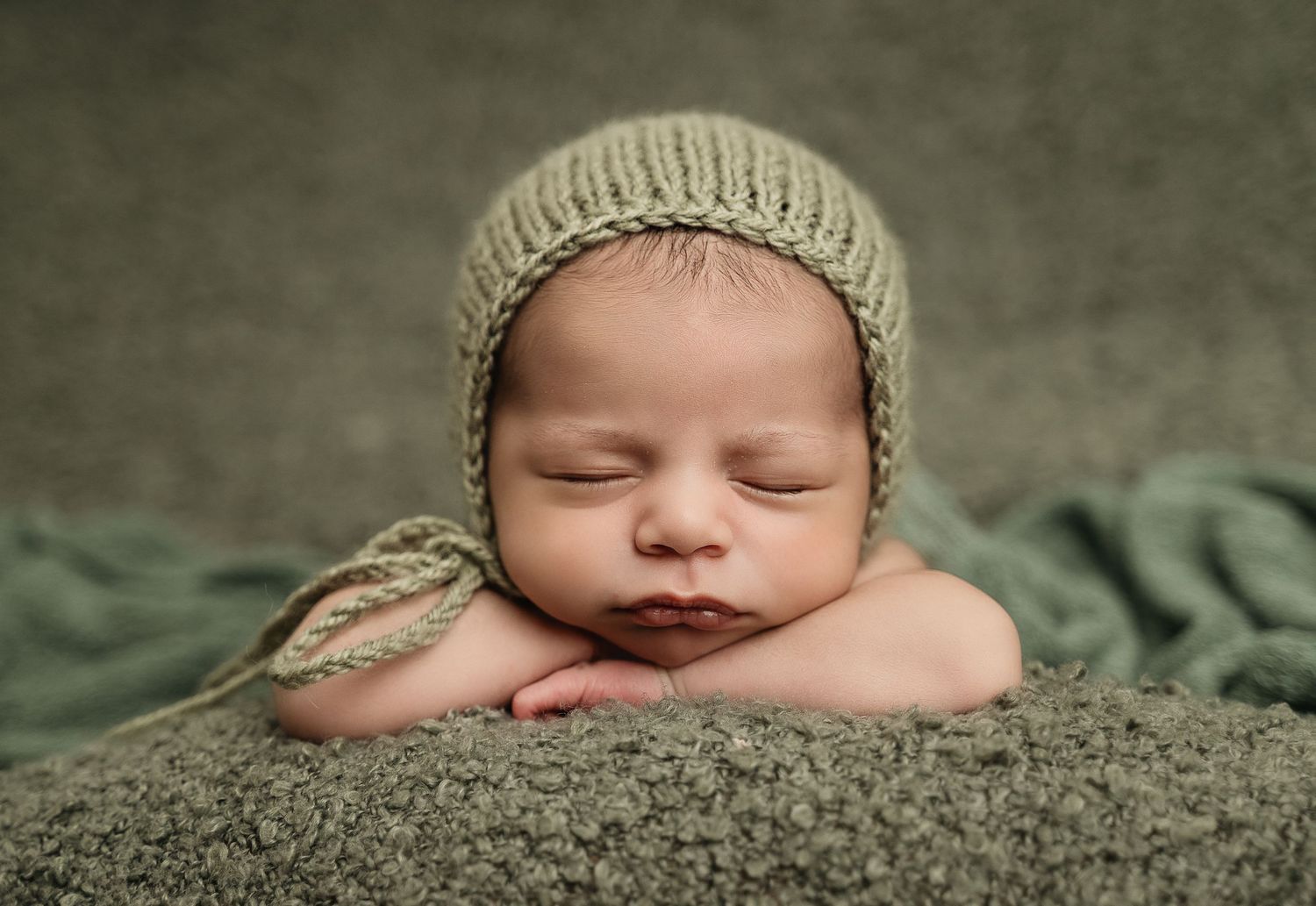 Sleeping newborn wearing knit beanie against soft green textured backdrop.