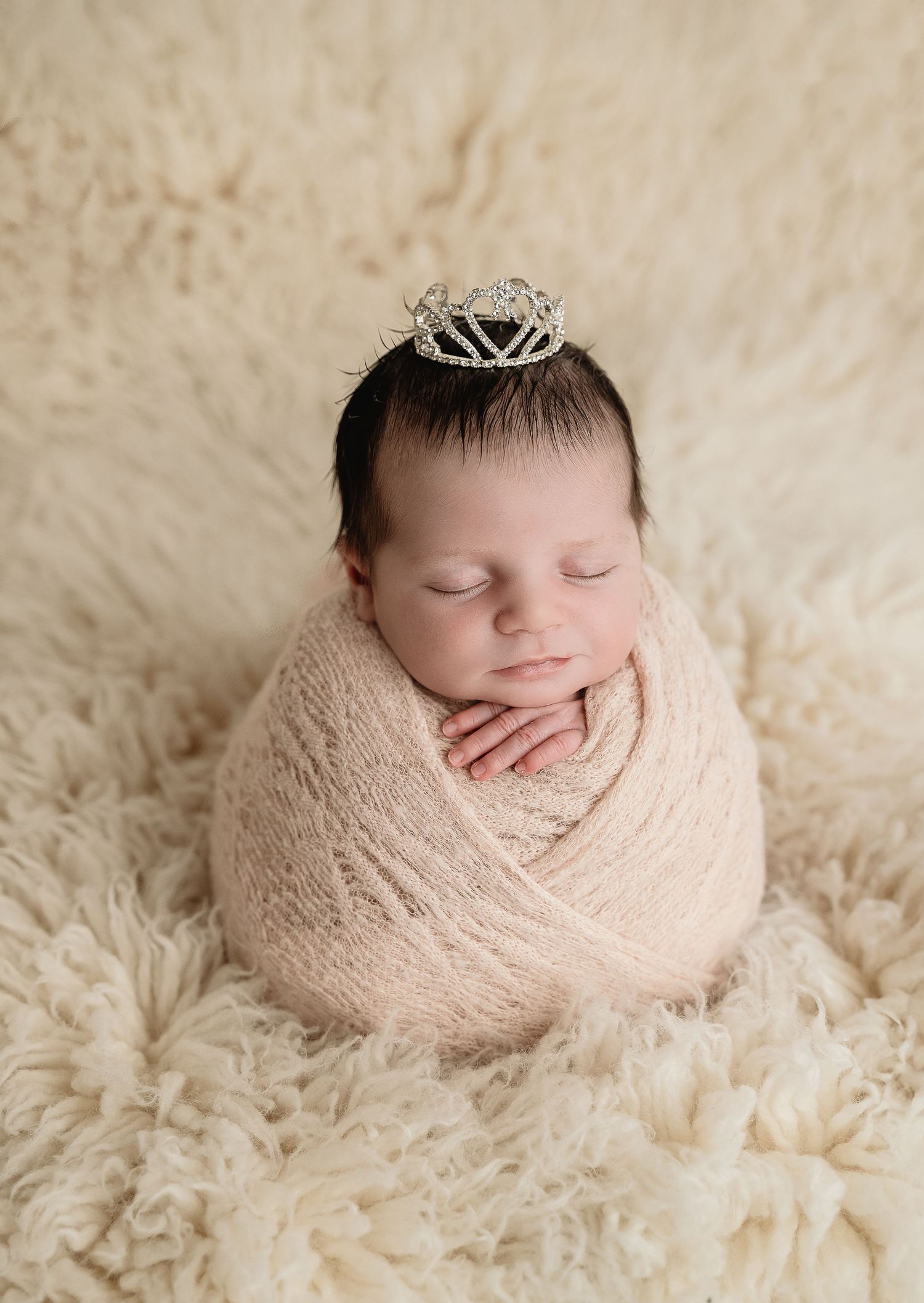 Peaceful sleeping newborn wrapped in beige fabric on fluffy cream textured backdrop.