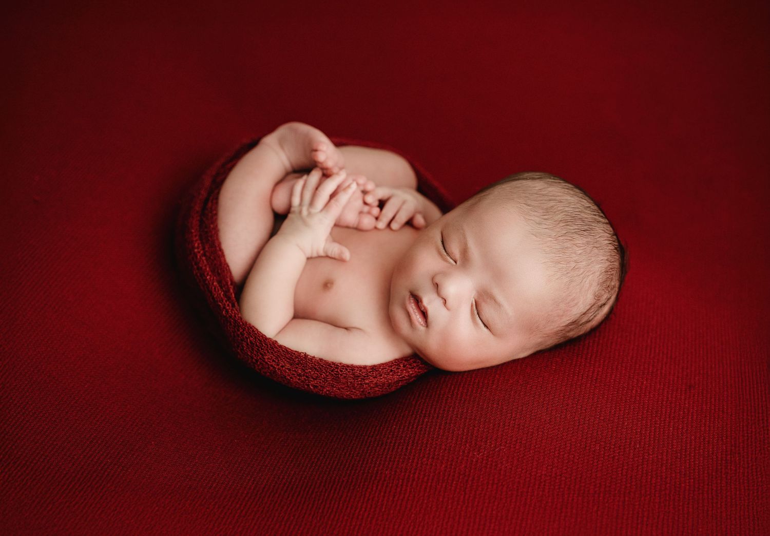 Newborn baby sleeping peacefully on soft burgundy red fabric in artistic portrait series.