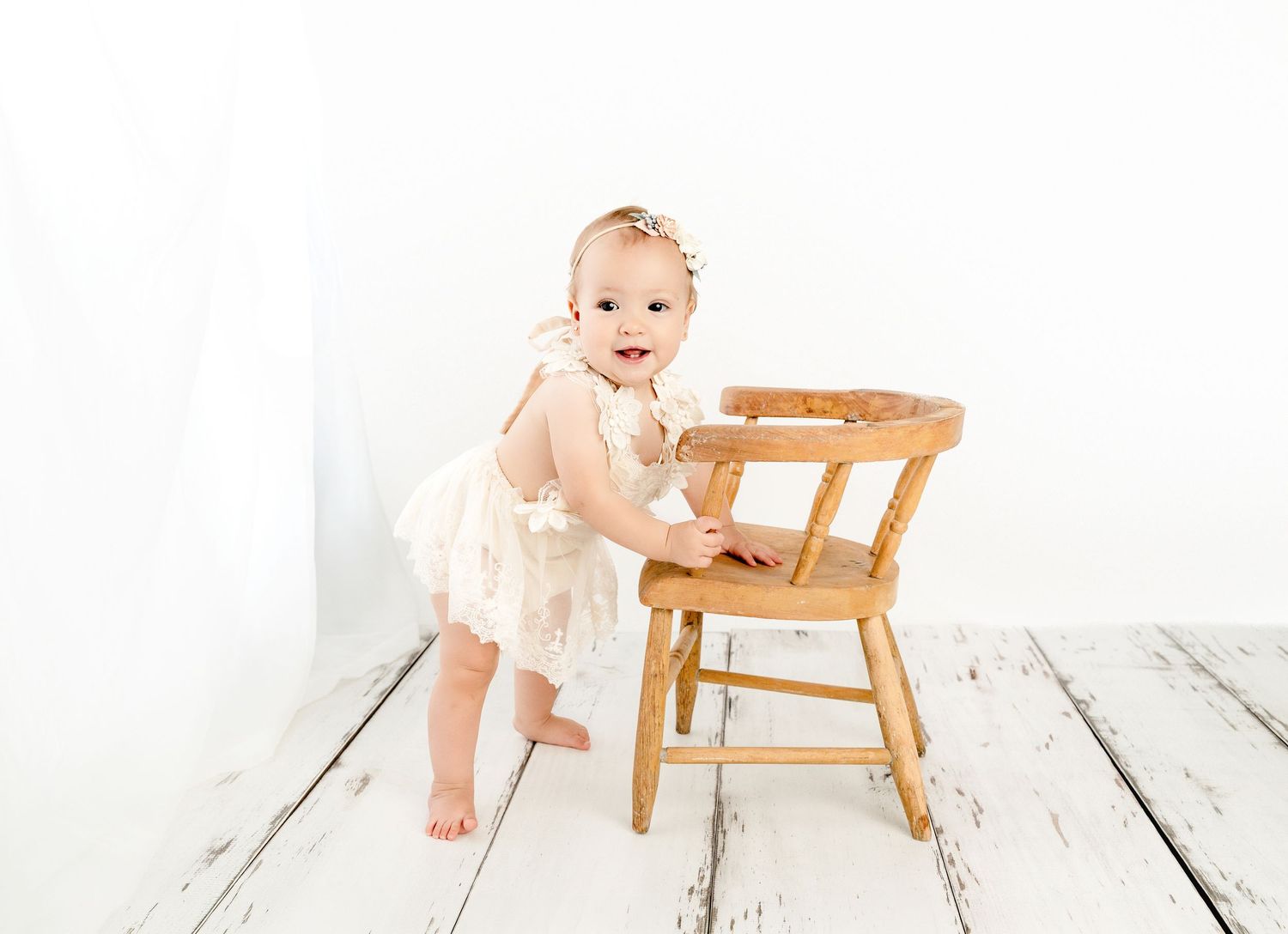 A baby girl in a lace dress stands holding a wooden chair on white floors.