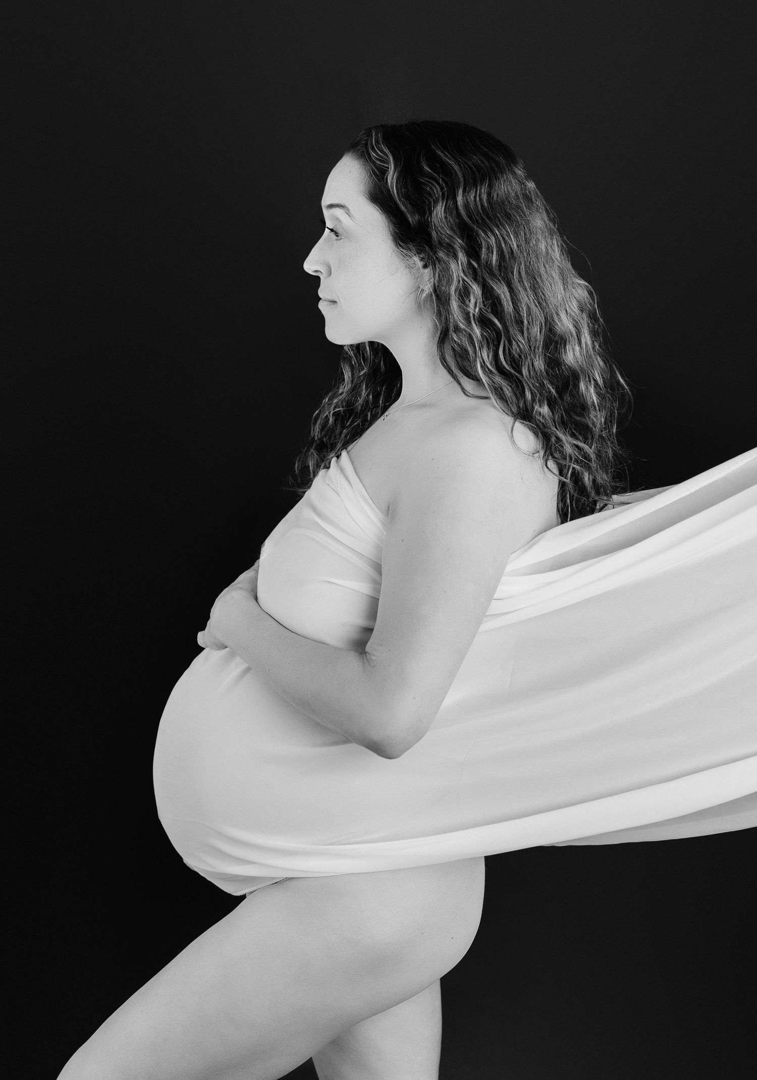 Black and white maternity photo of a pregnant woman with curly hair holding her belly, draped in flowing white fabric.