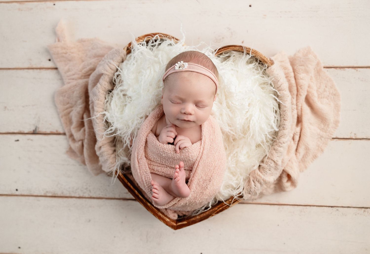 A baby wearing a white knit bonnet lies on a light wood plank background with soft pink accessories.