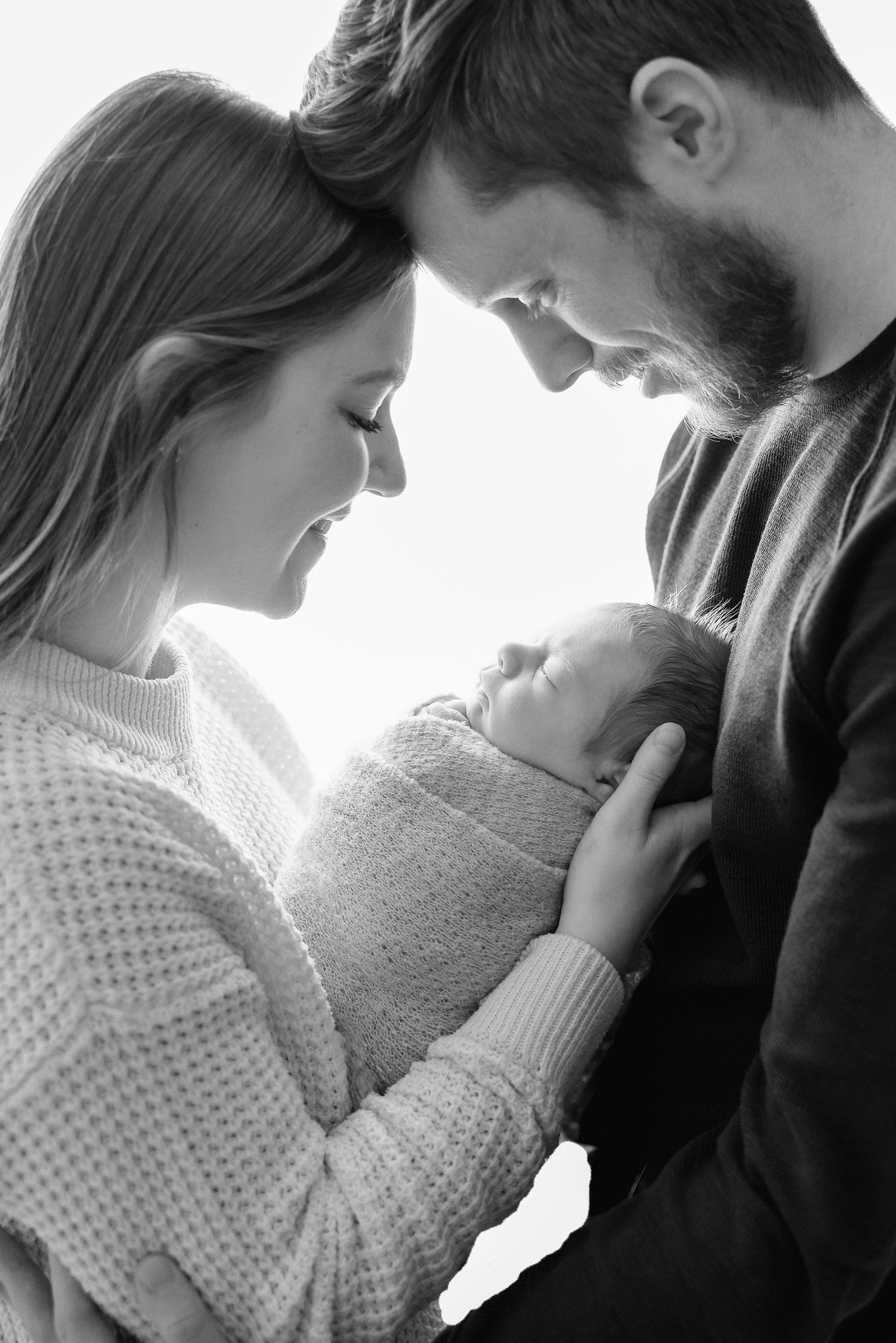 Intimate black and white portrait of parents cradling their newborn wrapped in soft blanket.