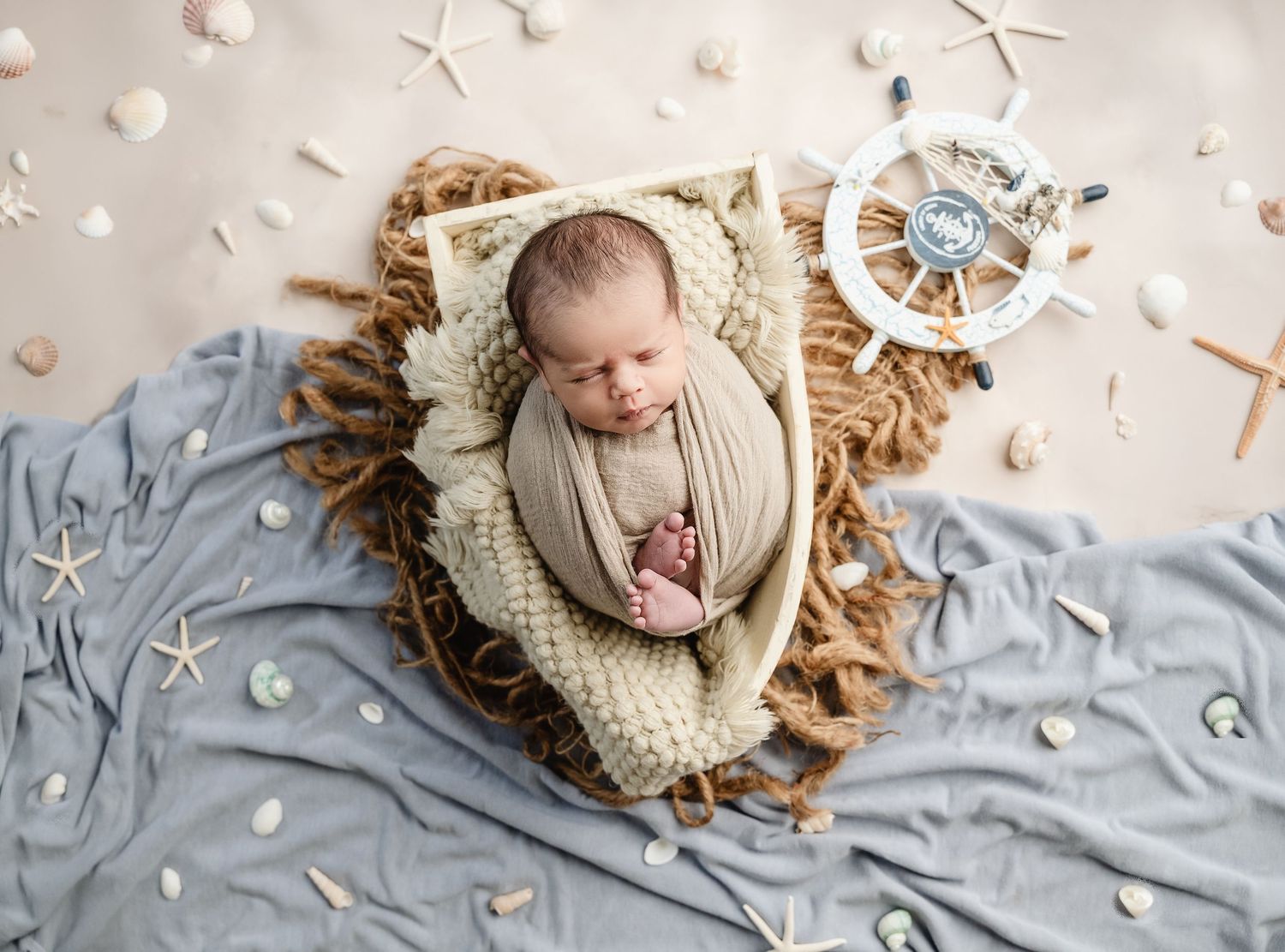 Newborn baby wrapped in cream swaddle sleeping in a rustic boat prop surrounded by seashells and starfish.