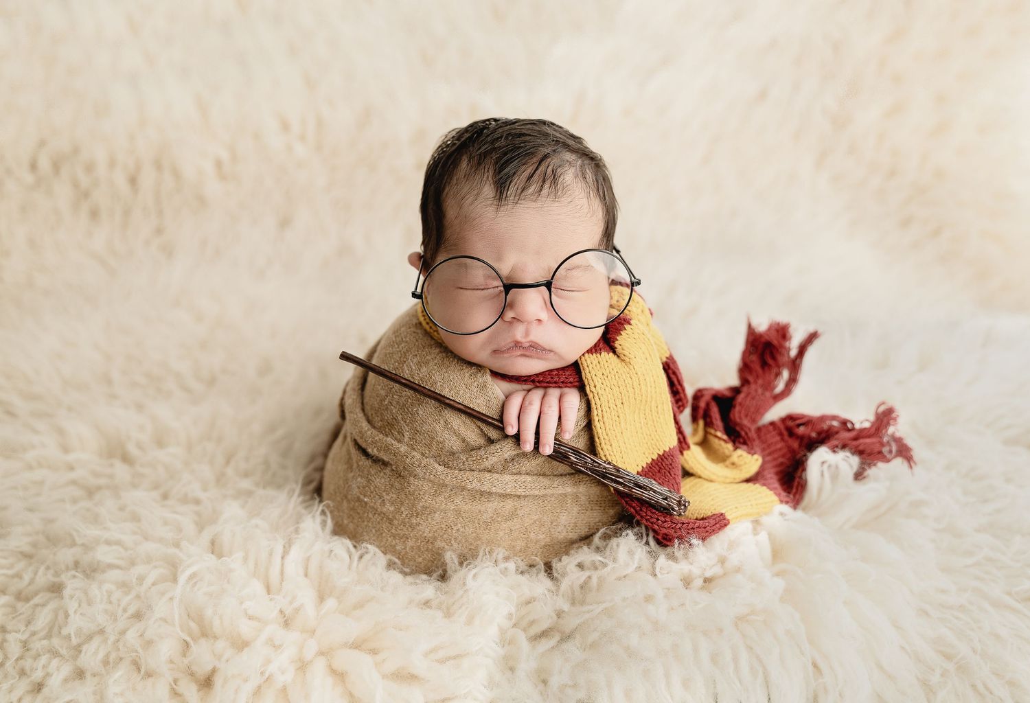 A baby in a tan wrap lies on a cream colored carpet with burgundy floral accents in a professional studio setting. Newborn session Denver