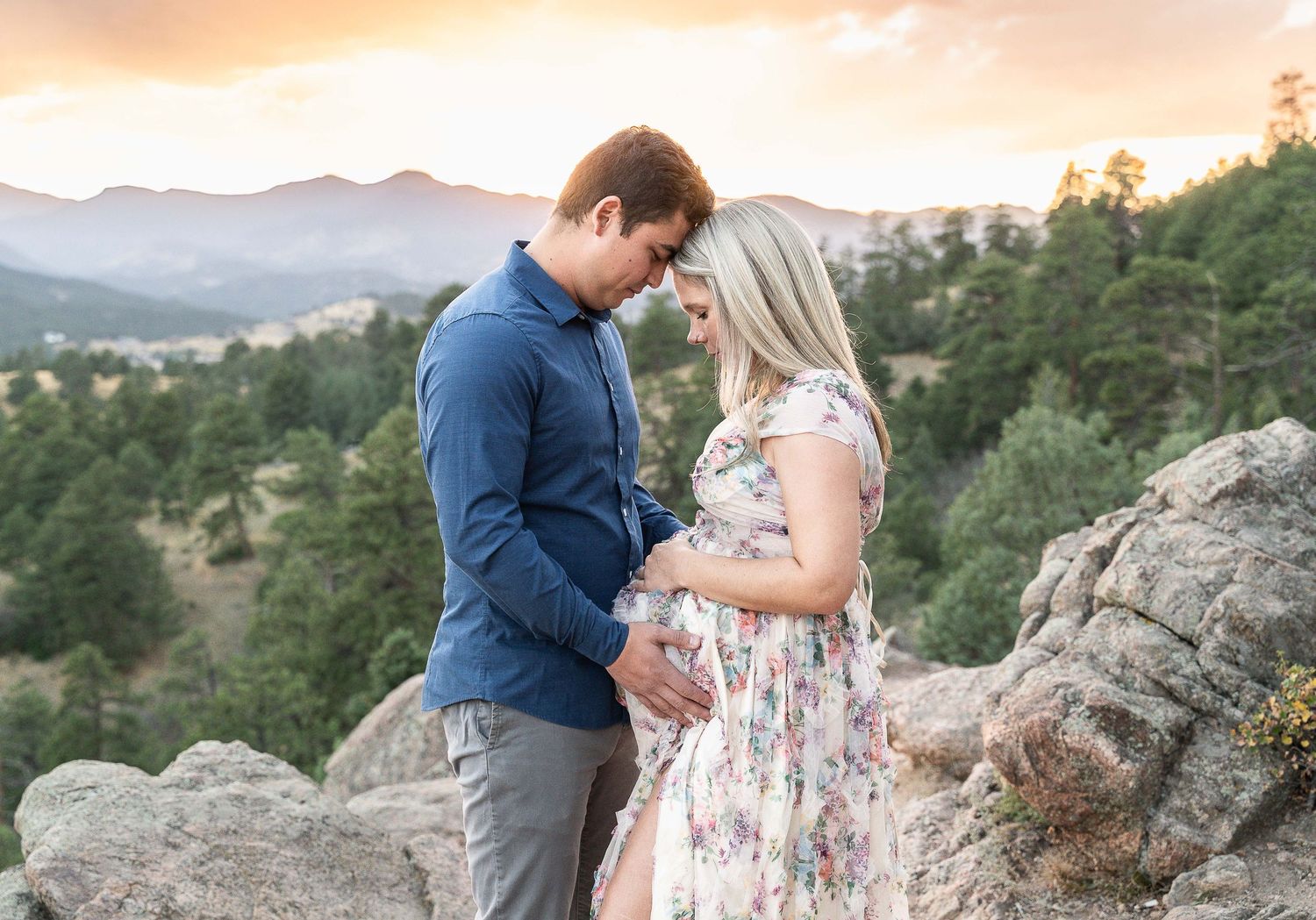 Two people share an intimate moment on a rocky overlook with mountains and evergreen forest at sunset.
