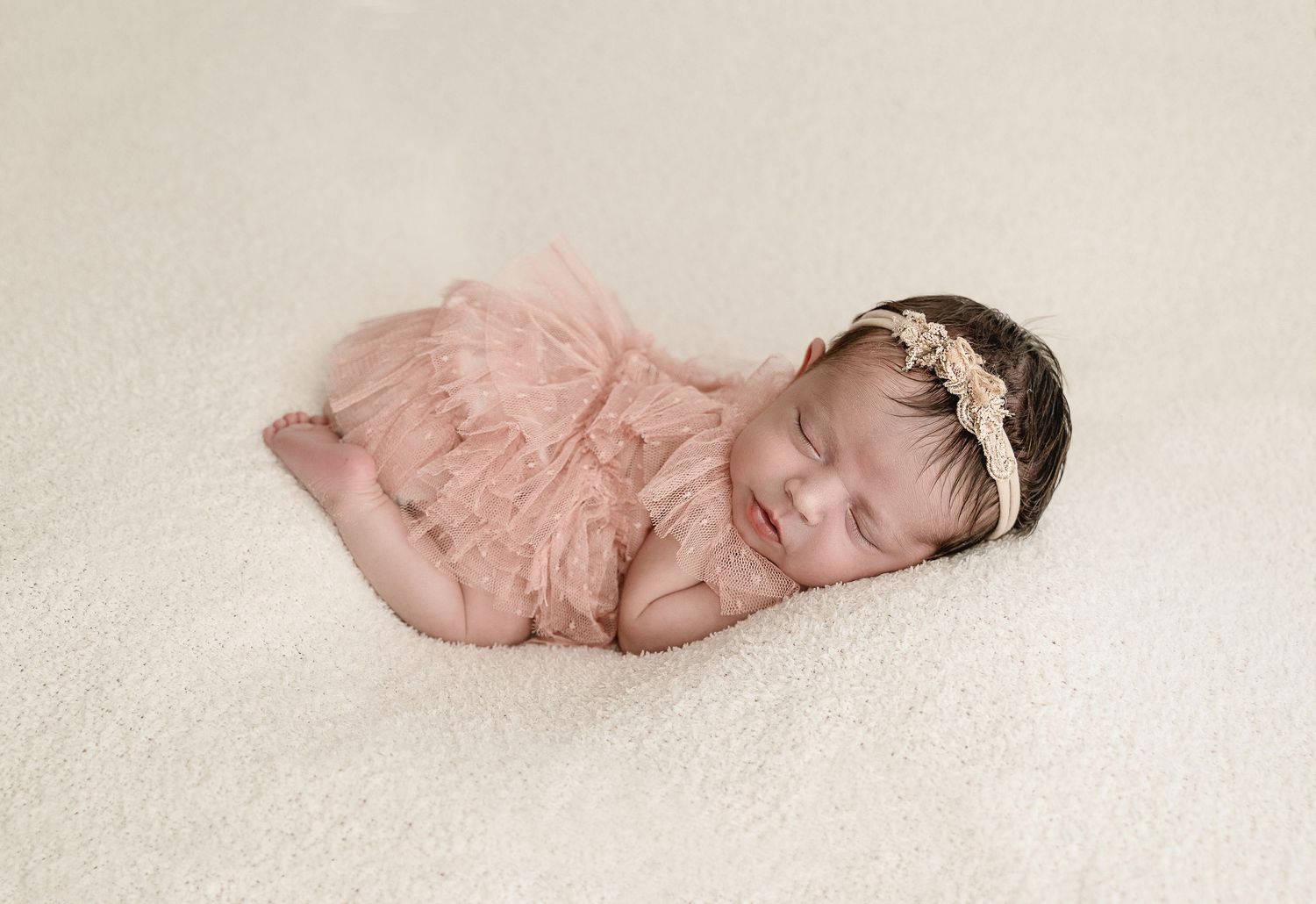 Sleeping newborn baby wearing pink lace wrap and floral headband lies on cream colored backdrop.