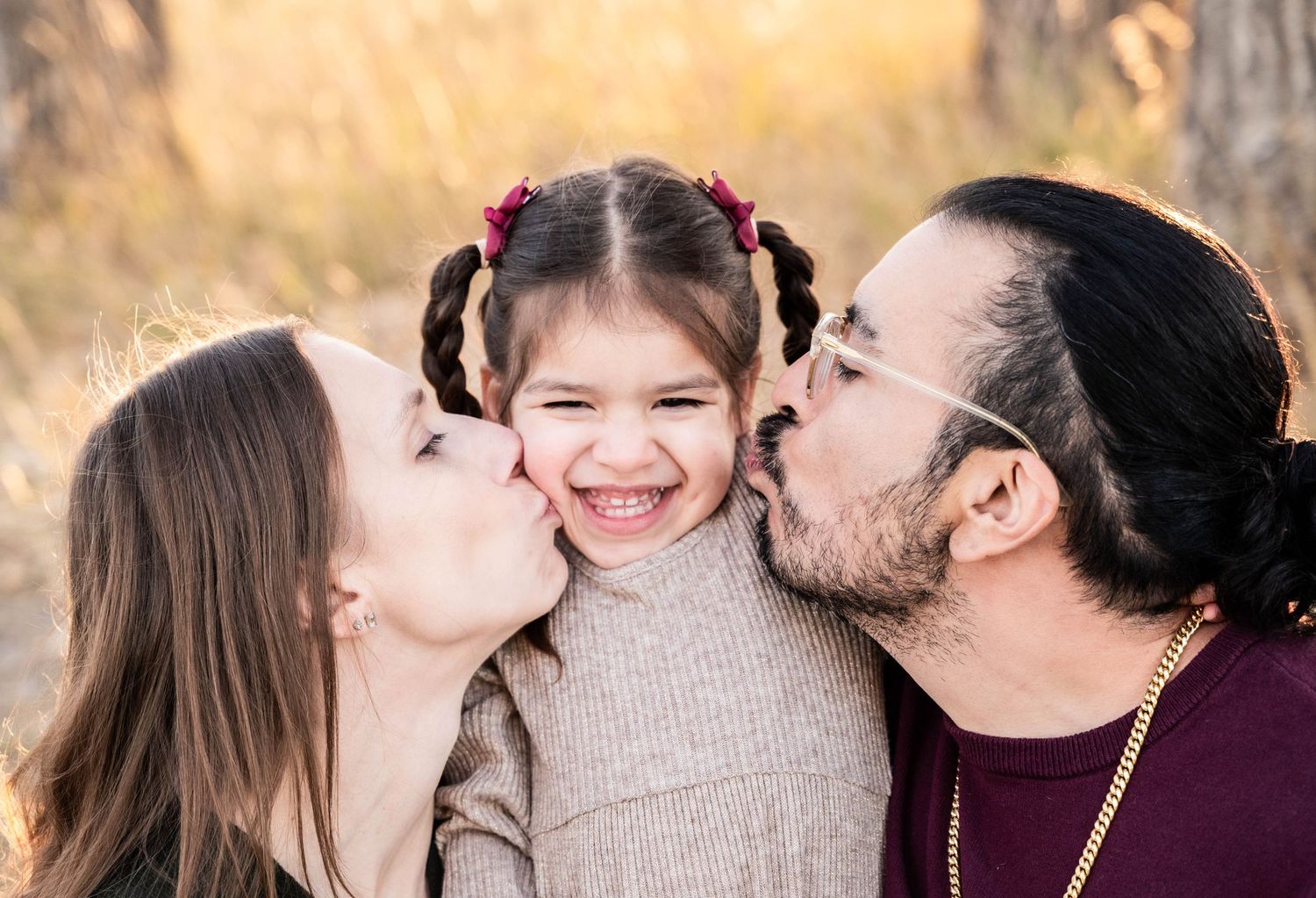 Parents kiss their delighted child on both cheeks during a warm sunset photoshoot in nature.
