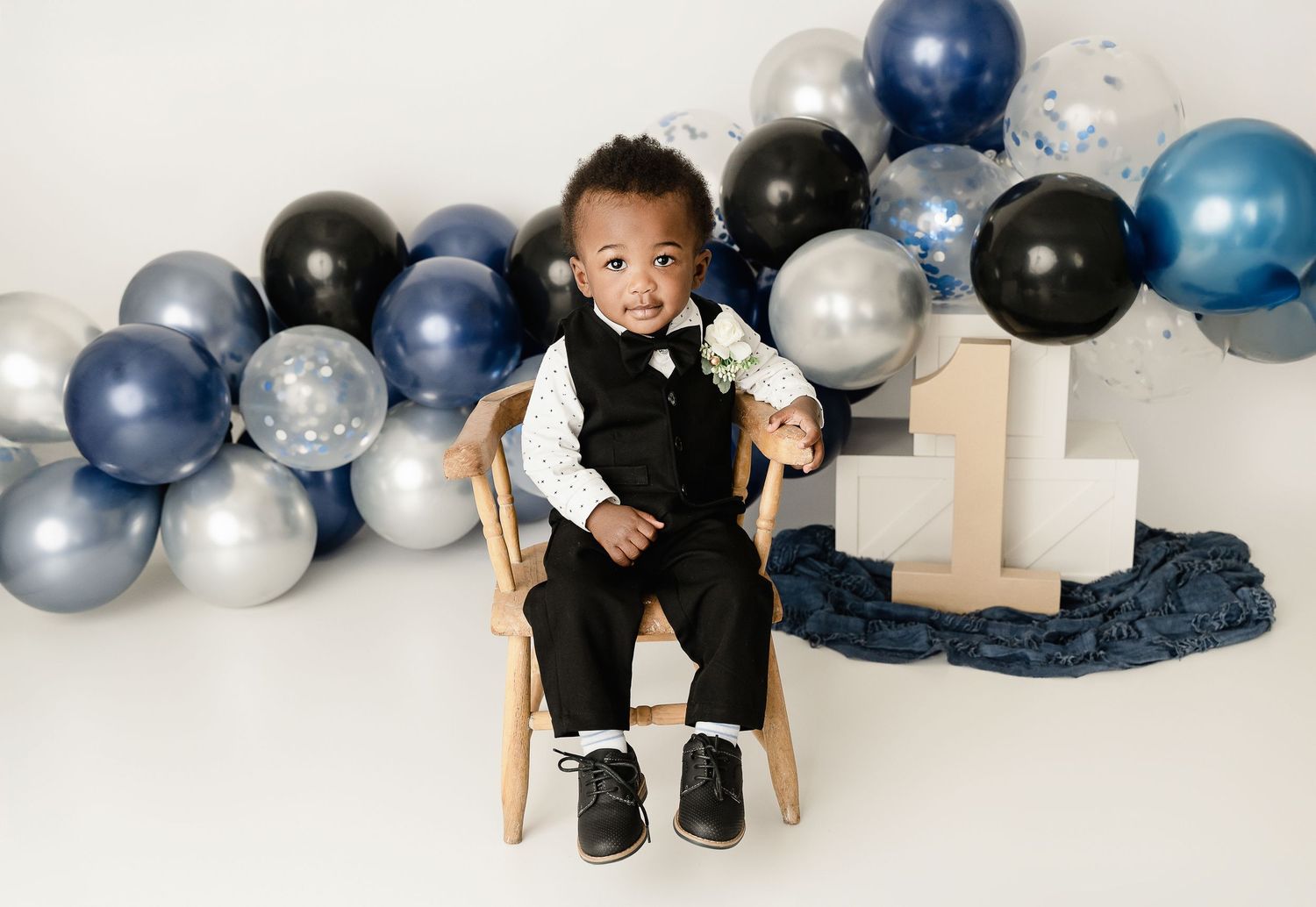 First birthday setup with navy and silver balloon arch and toddler dressed in formal attire sitting in wooden chair.