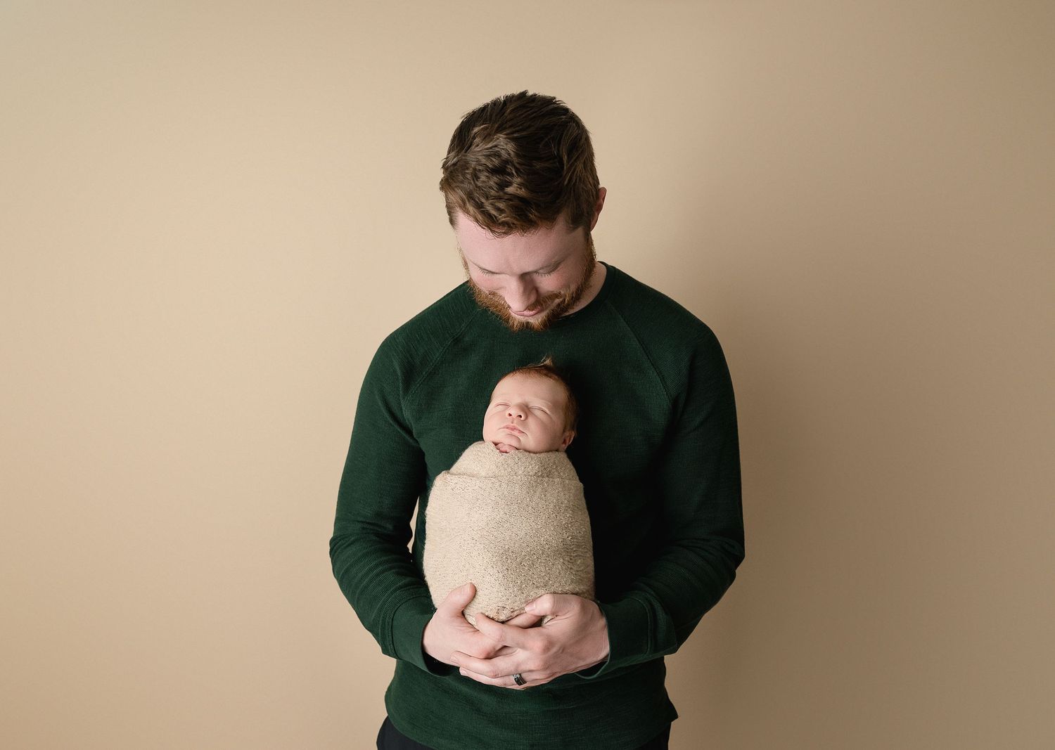 Father in dark green sweater tenderly holding sleeping newborn against beige background.