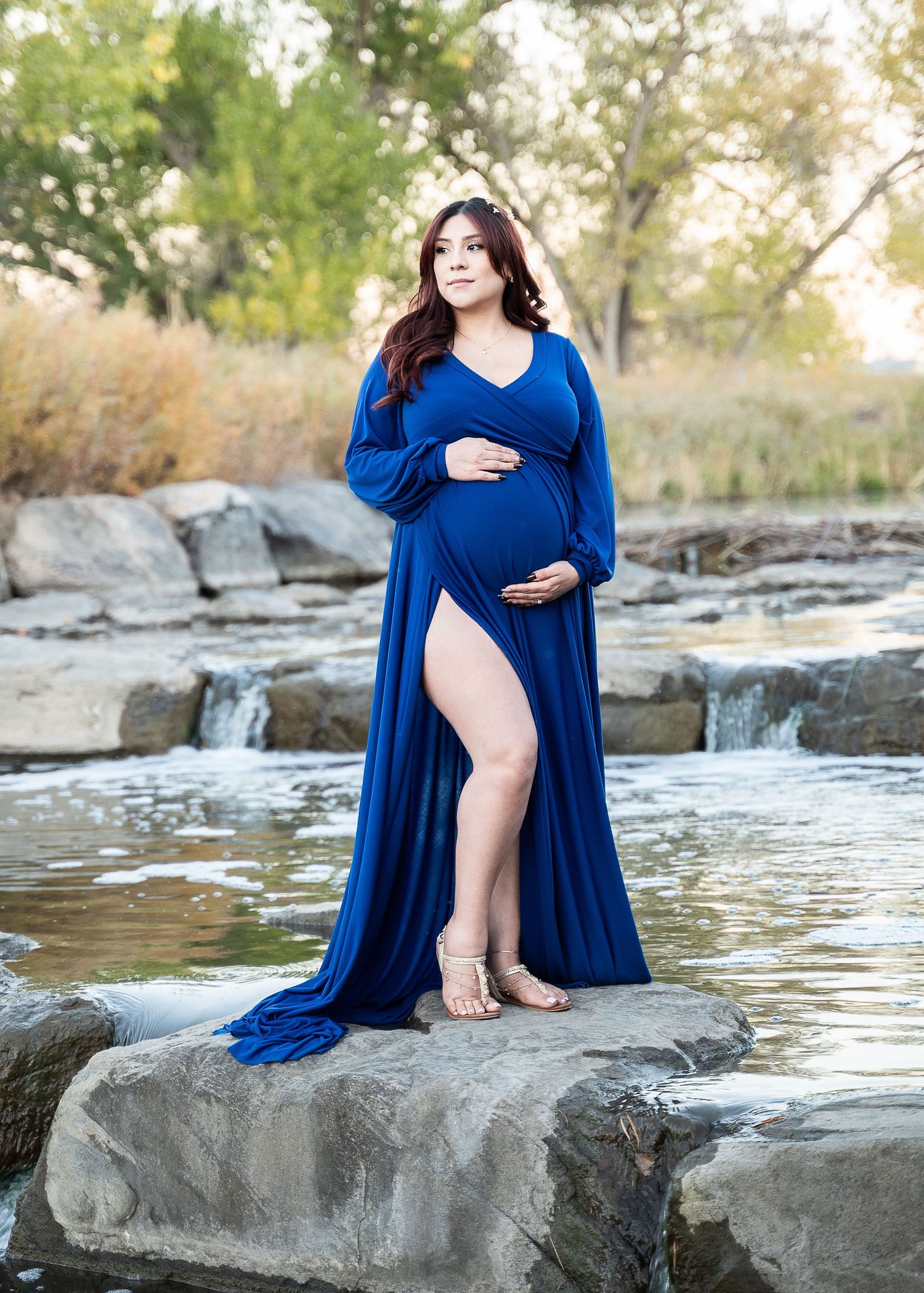 Expectant mother in flowing royal blue dress poses gracefully on rocks near a waterfall.