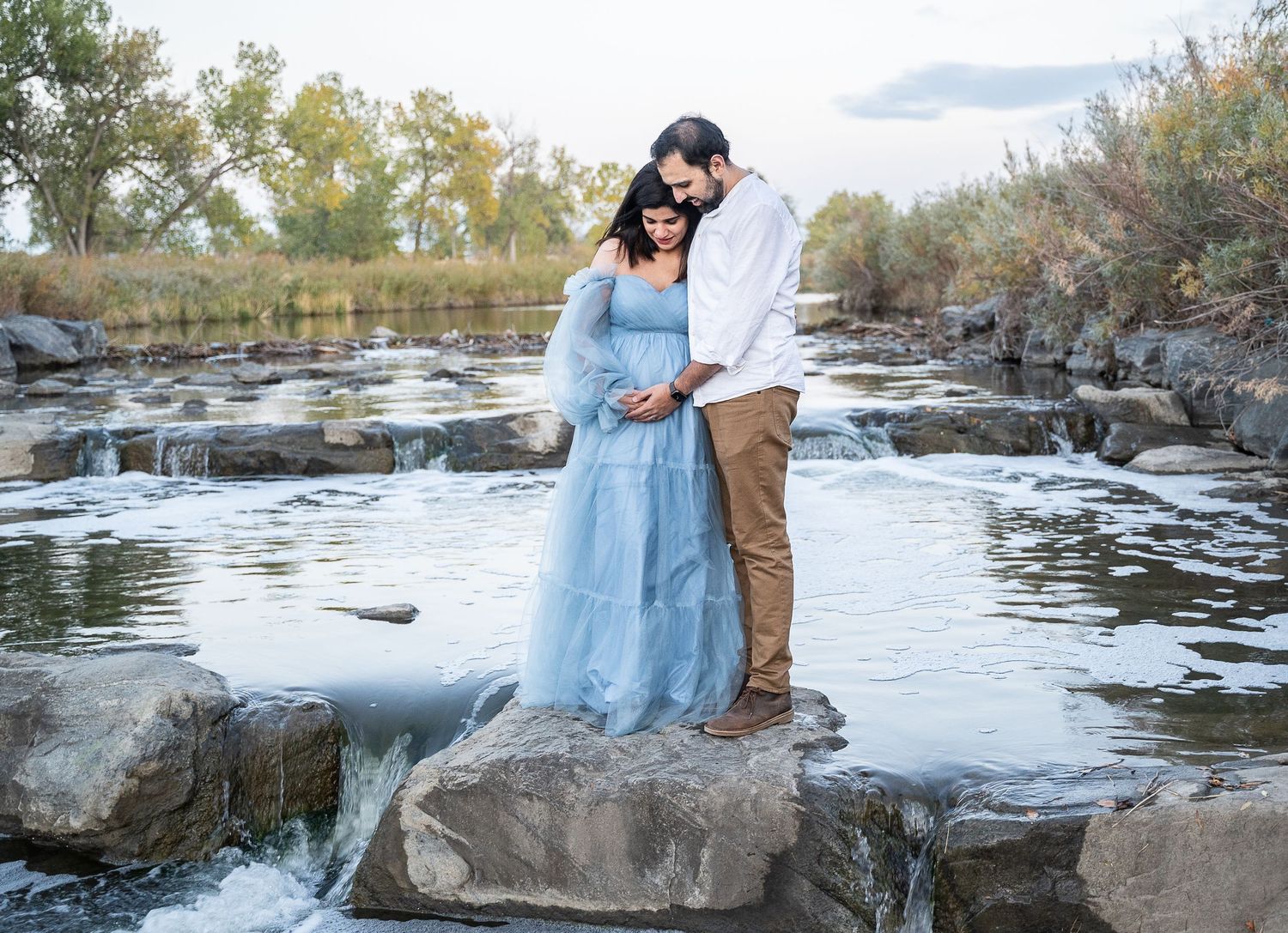 Romantic maternity photo of couple standing on rocks by flowing stream.