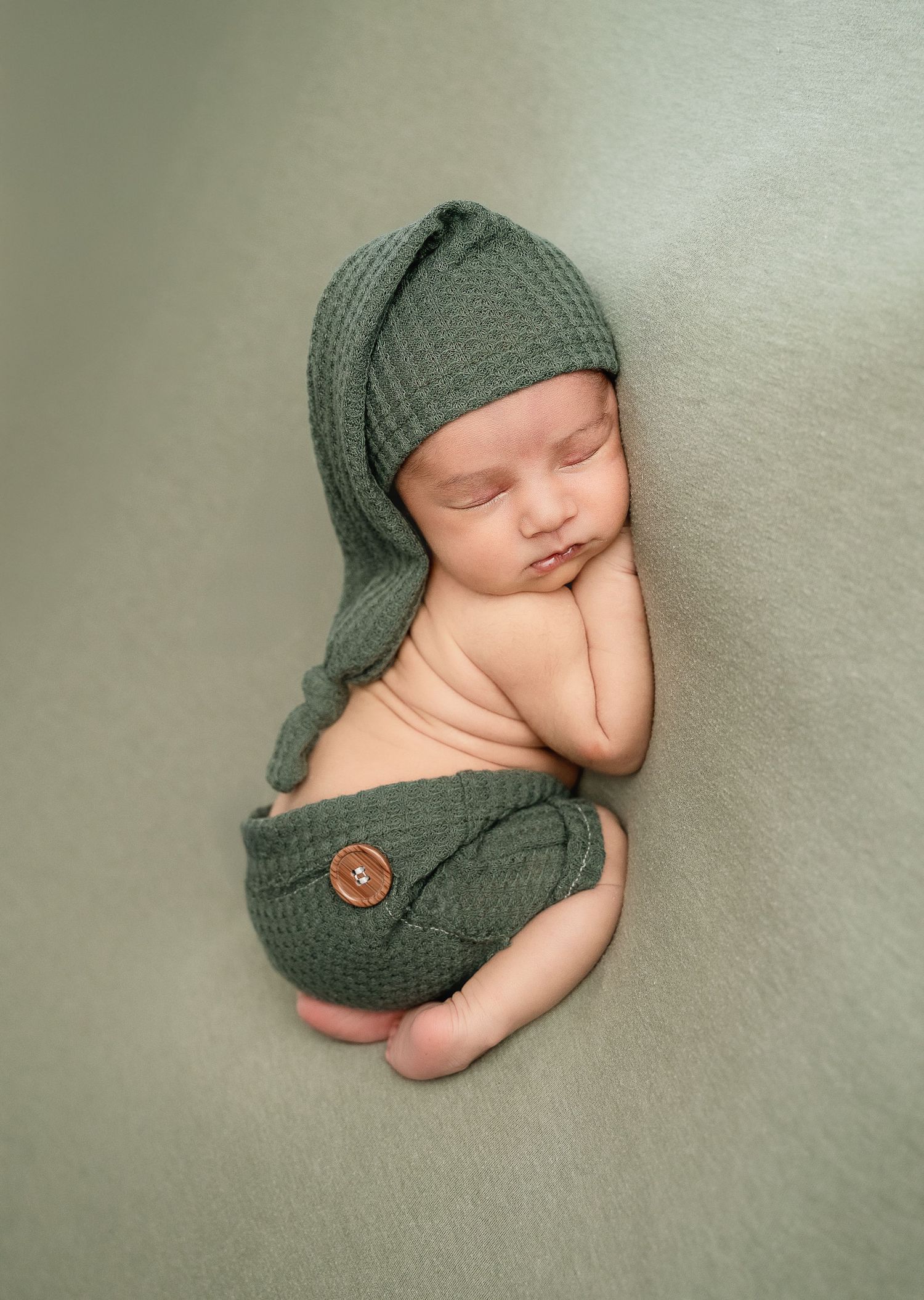 Sleeping newborn wearing green knit hat and bottoms posed against sage green background.