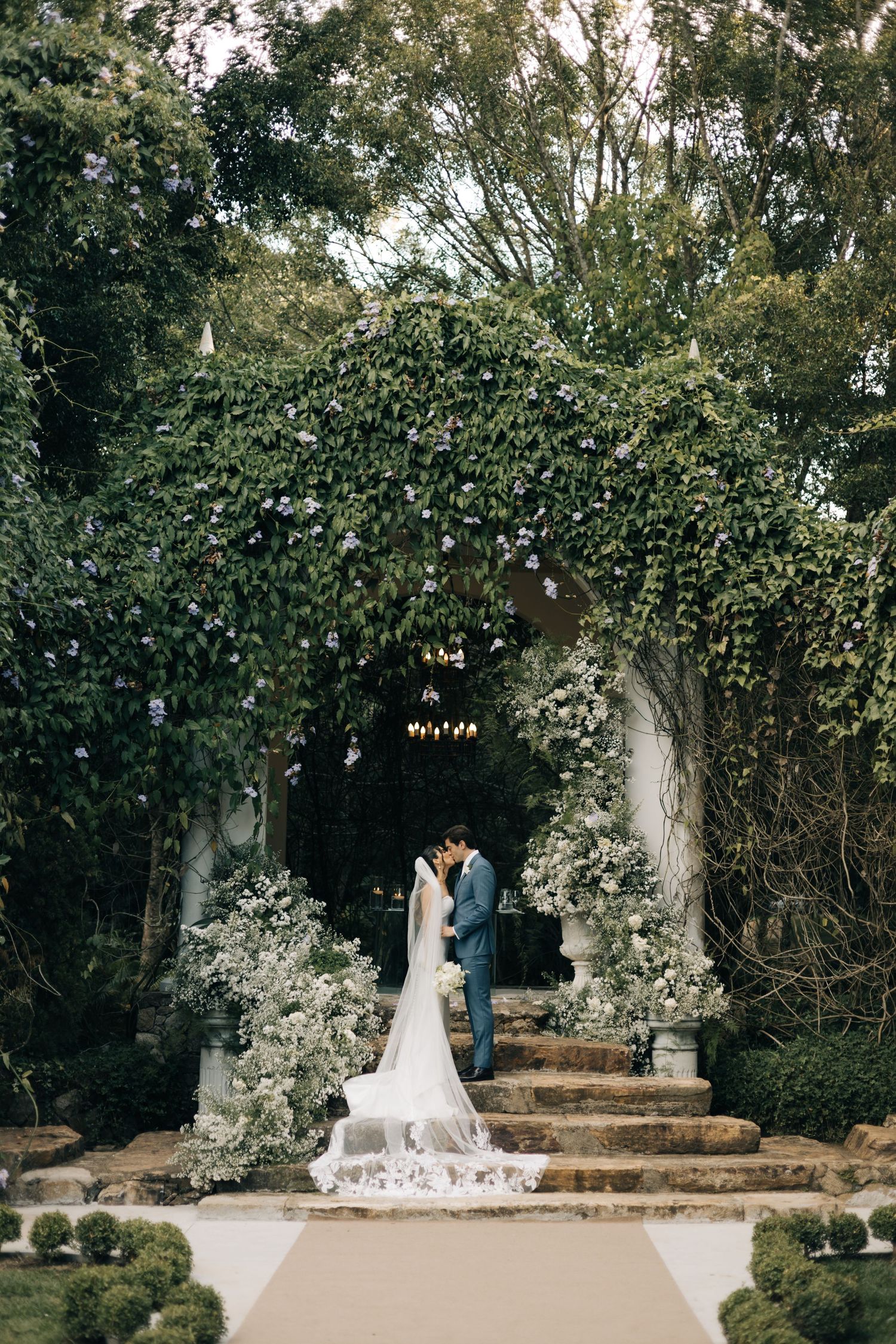 Bride and groom kissing under an ivy-covered archway adorned with white flowers at an elegant outdoor wedding ceremony.