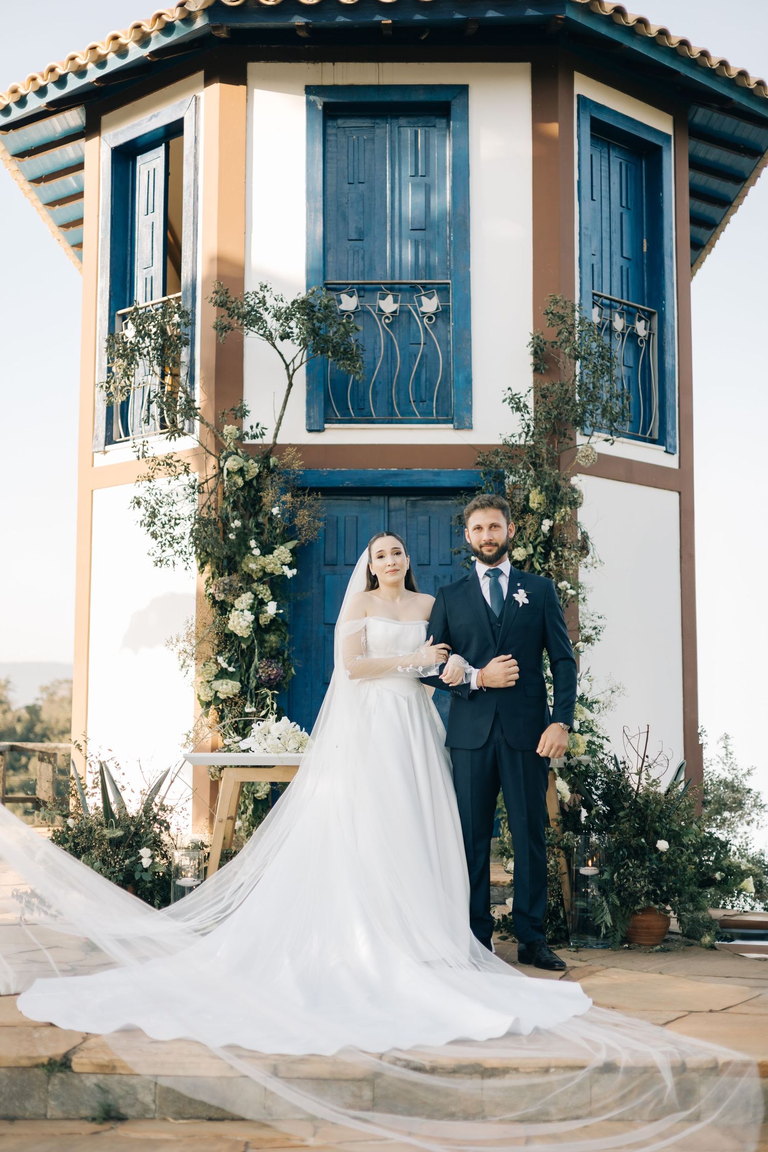 A wedding couple poses in front of a Mediterranean-style tower with blue doors and climbing vines.